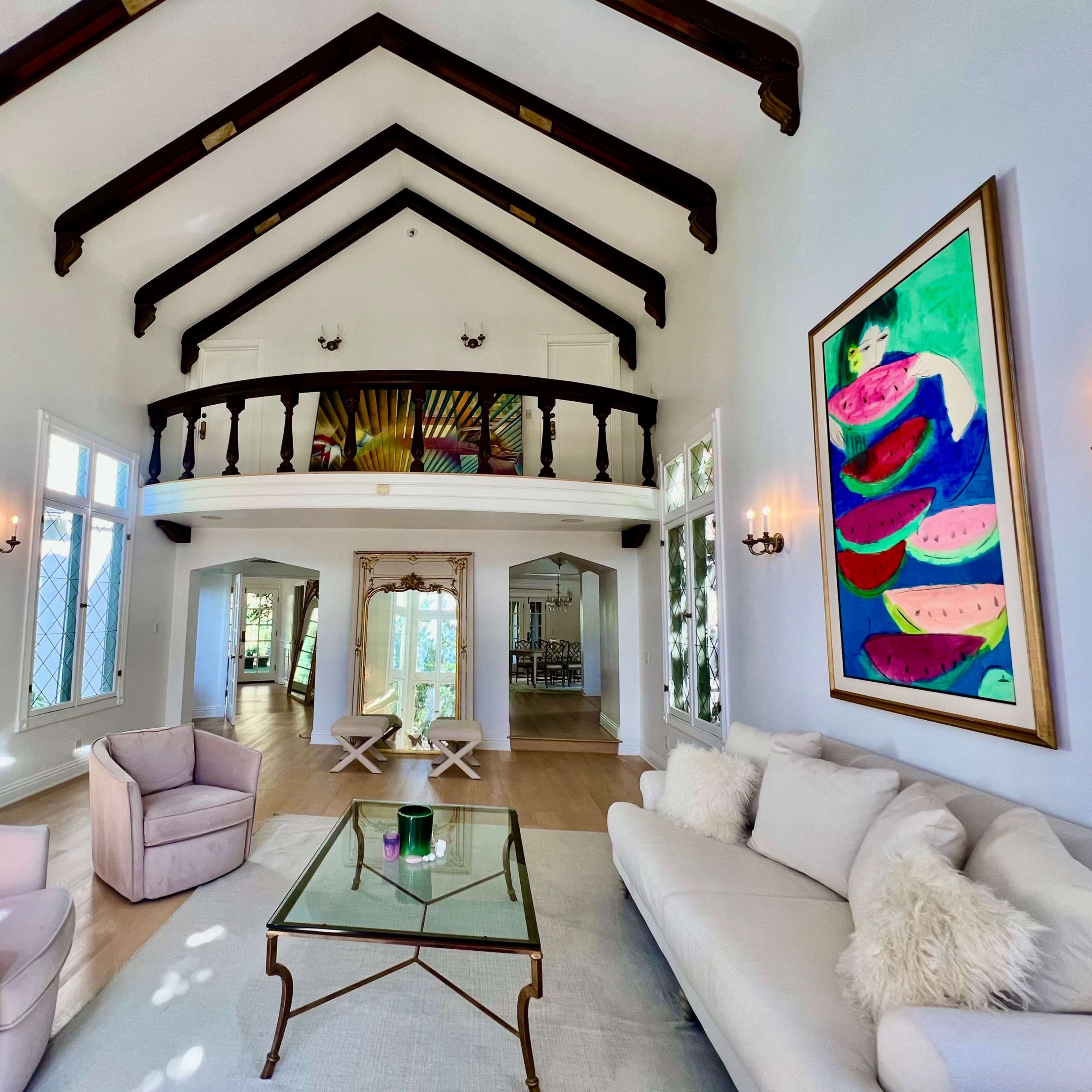 A bright living room with high vaulted ceilings, large windows, and a comfortable seating area featuring a sofa and armchairs, complemented by colorful artwork on the walls.