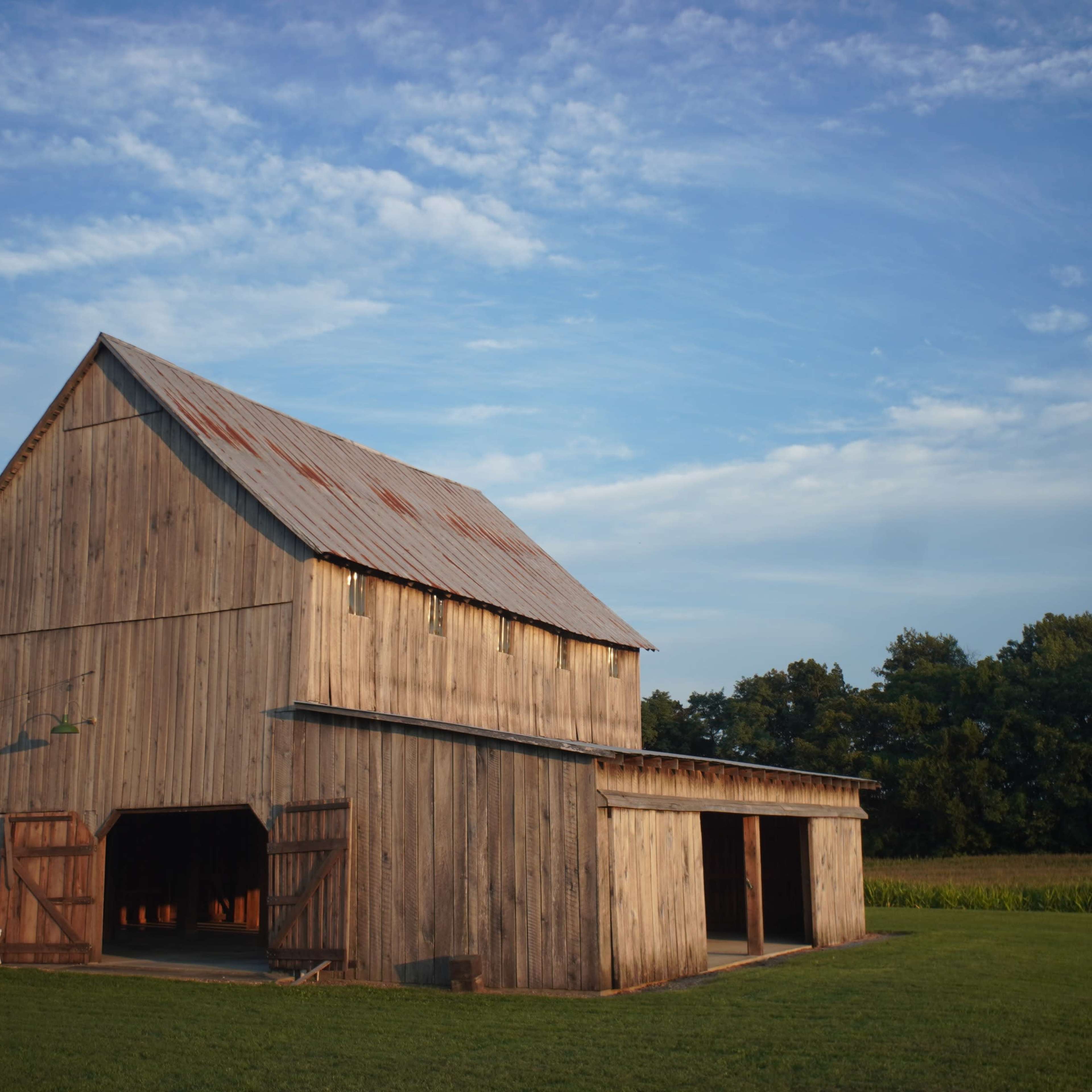 A wooden barn with a rusty metal roof stands in a field against a blue sky with scattered clouds.