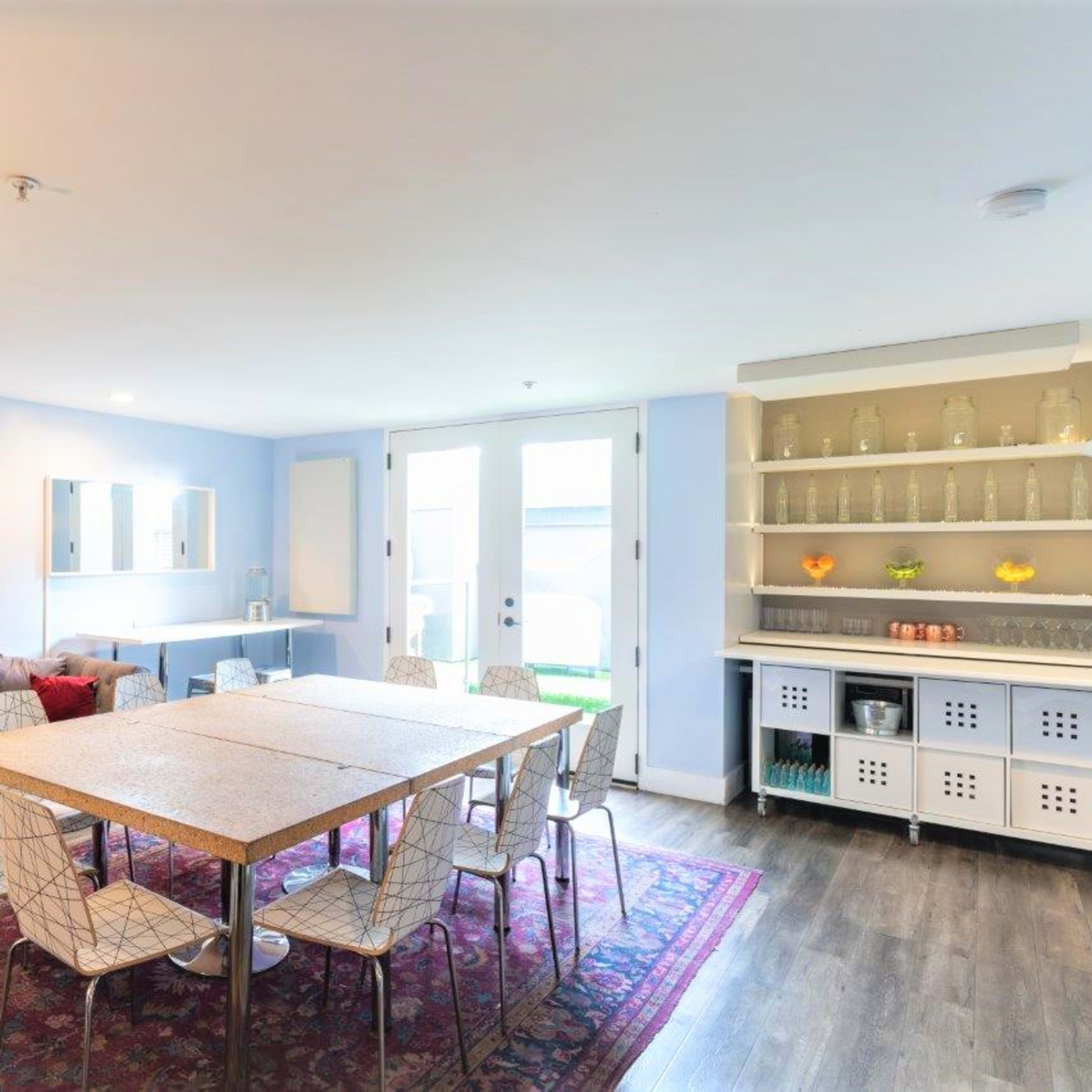 A dining area features a large table surrounded by chairs, with a sofa and a shelf filled with glassware in a well-lit, modern room.