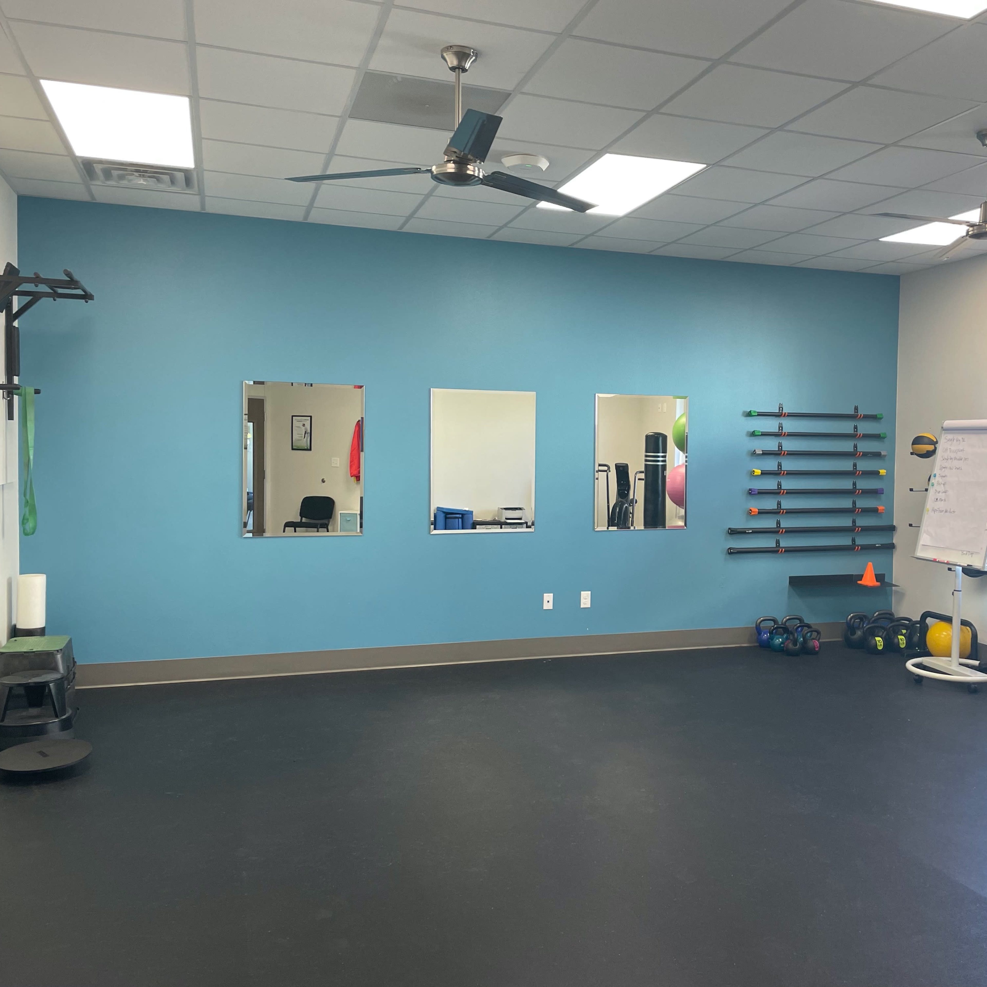 A well-lit gym interior featuring exercise equipment, three mirrors on a blue wall, and a black rubber floor.