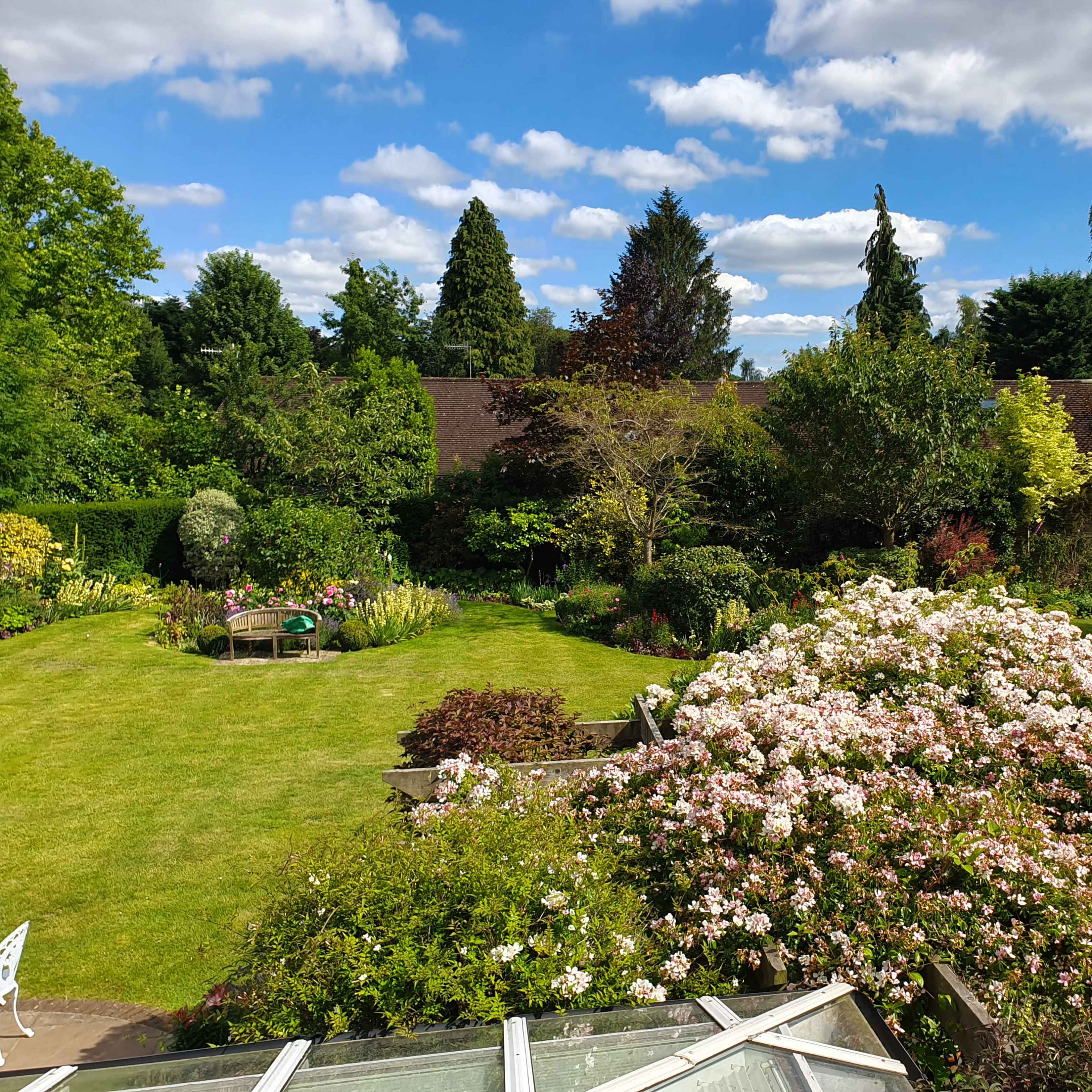 A well-maintained garden featuring a lush lawn, blooming flowers, and trees under a partly cloudy sky.