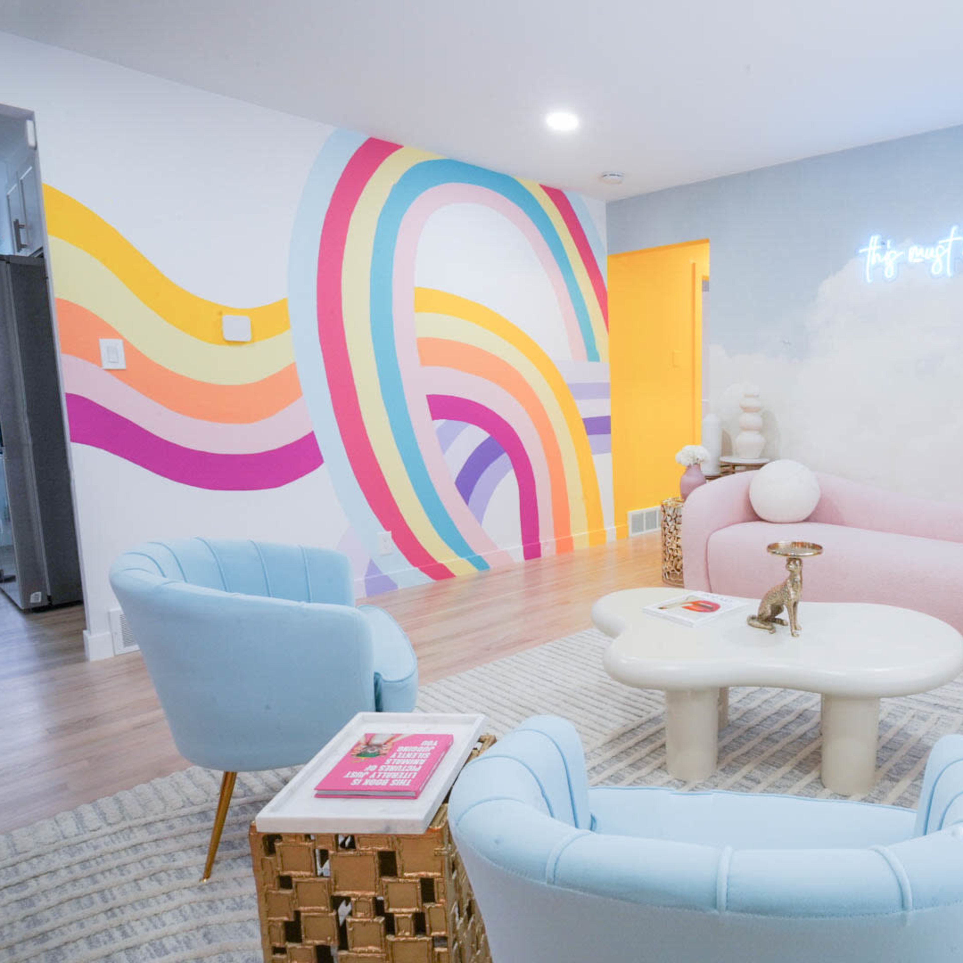 A brightly colored lounge area with pastel furniture, a rainbow mural on the wall, and a neon sign that reads "It's easy to take place."