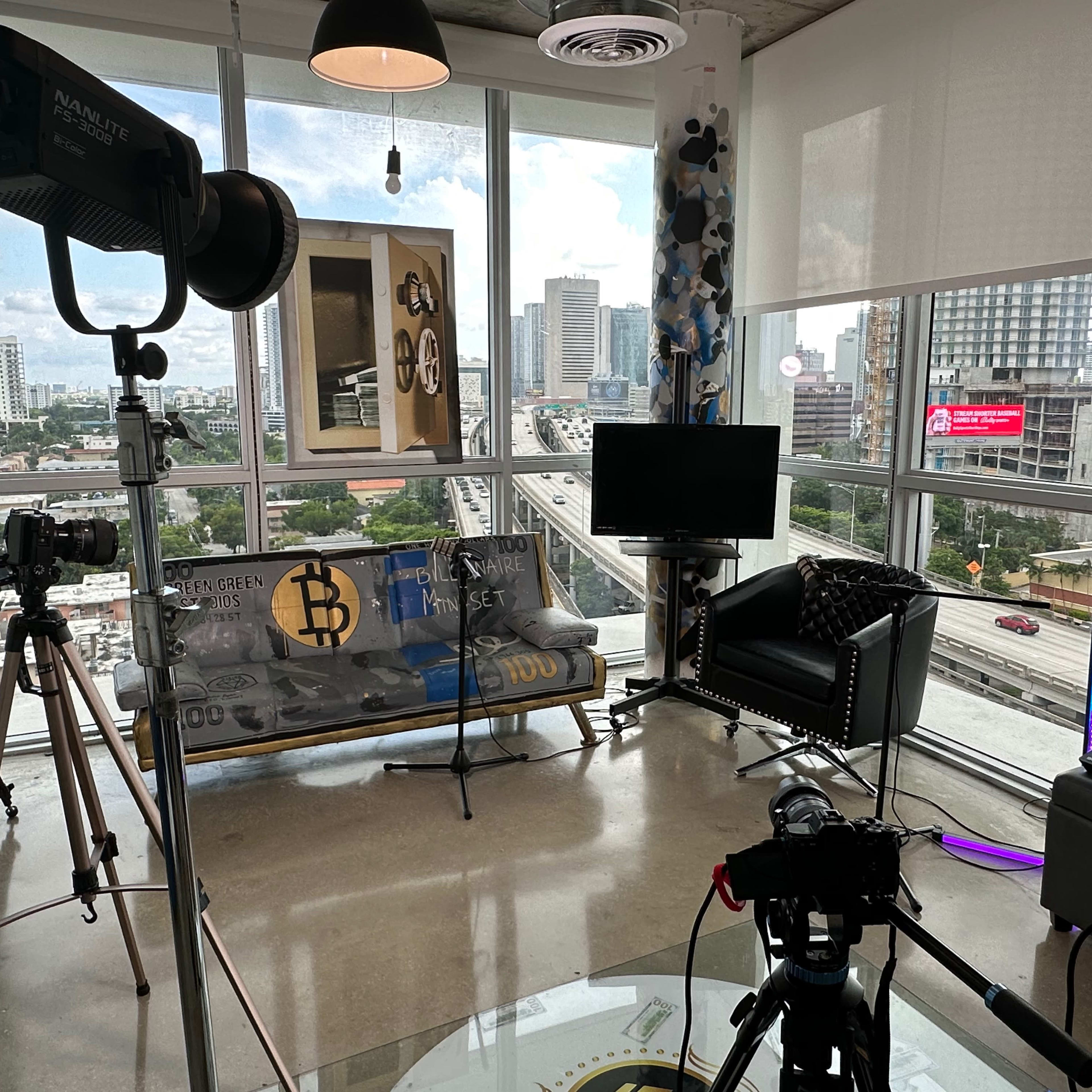 A modern studio space features a couch, television, and camera equipment, with large windows overlooking a city street.