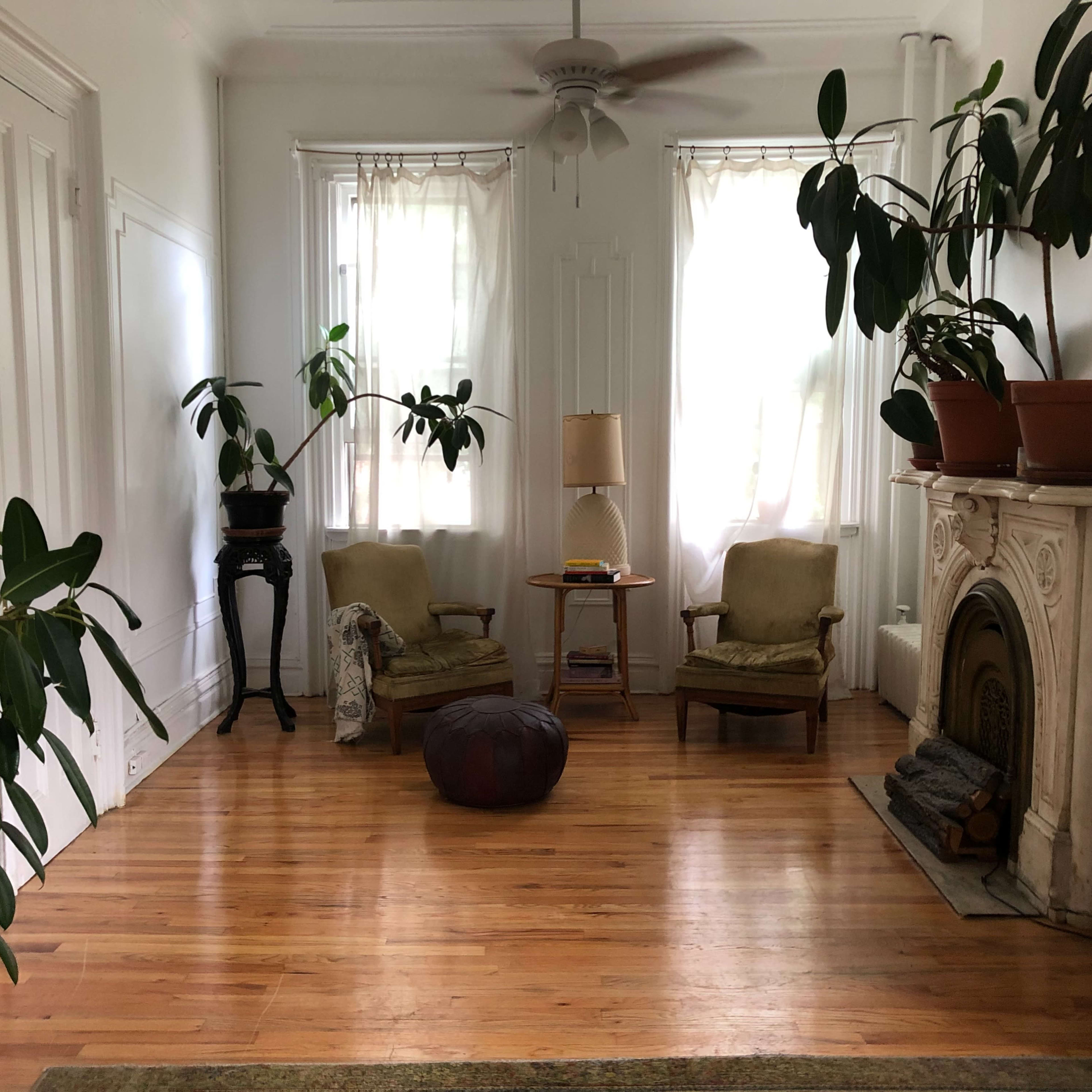 A spacious living room with two chairs, a small table, and several potted plants near a decorative fireplace.