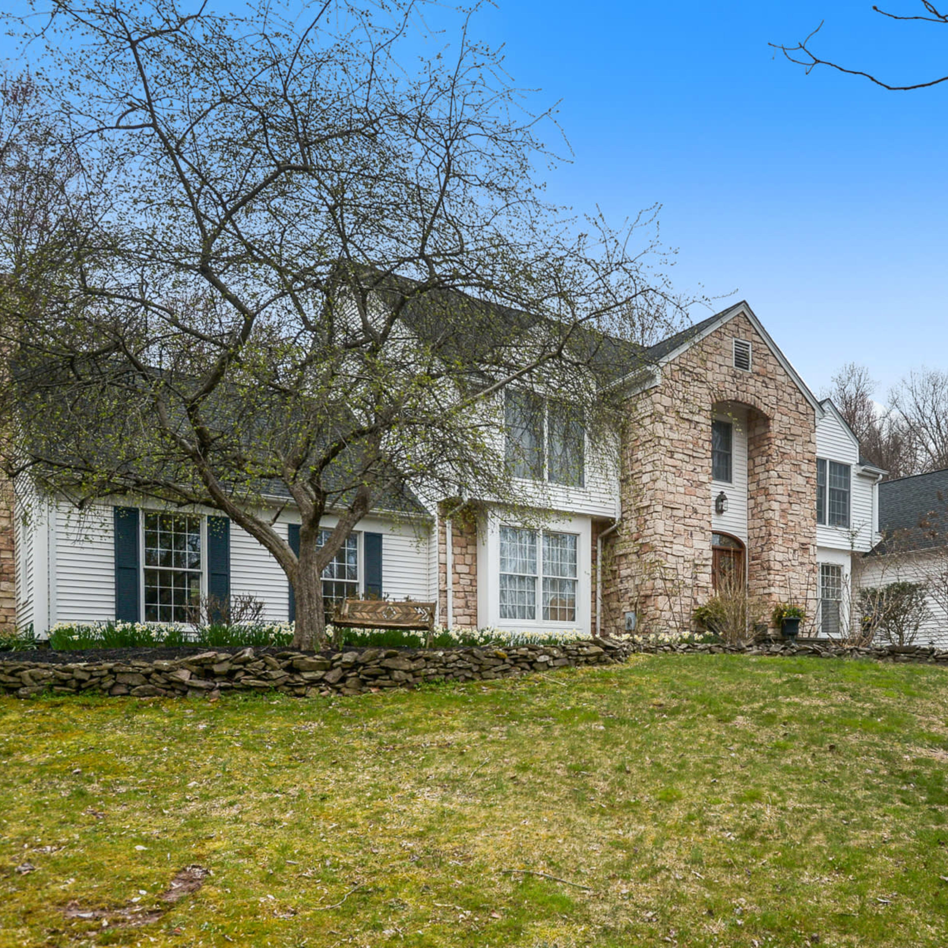 A large stone and siding house is set on a grassy lawn, surrounded by trees and shrubs.