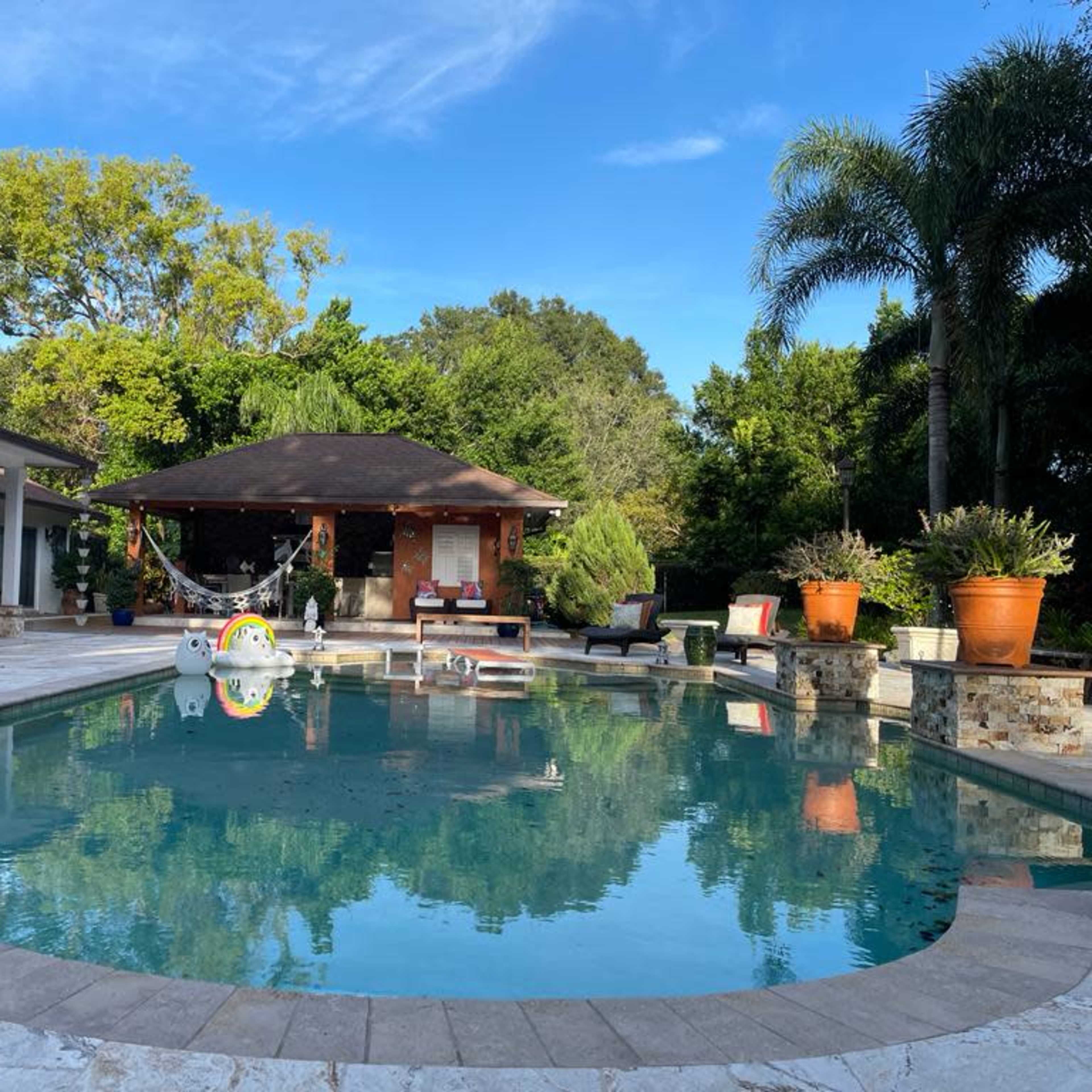A residential backyard with a large swimming pool surrounded by stone borders, a gazebo, potted plants, and sun loungers.