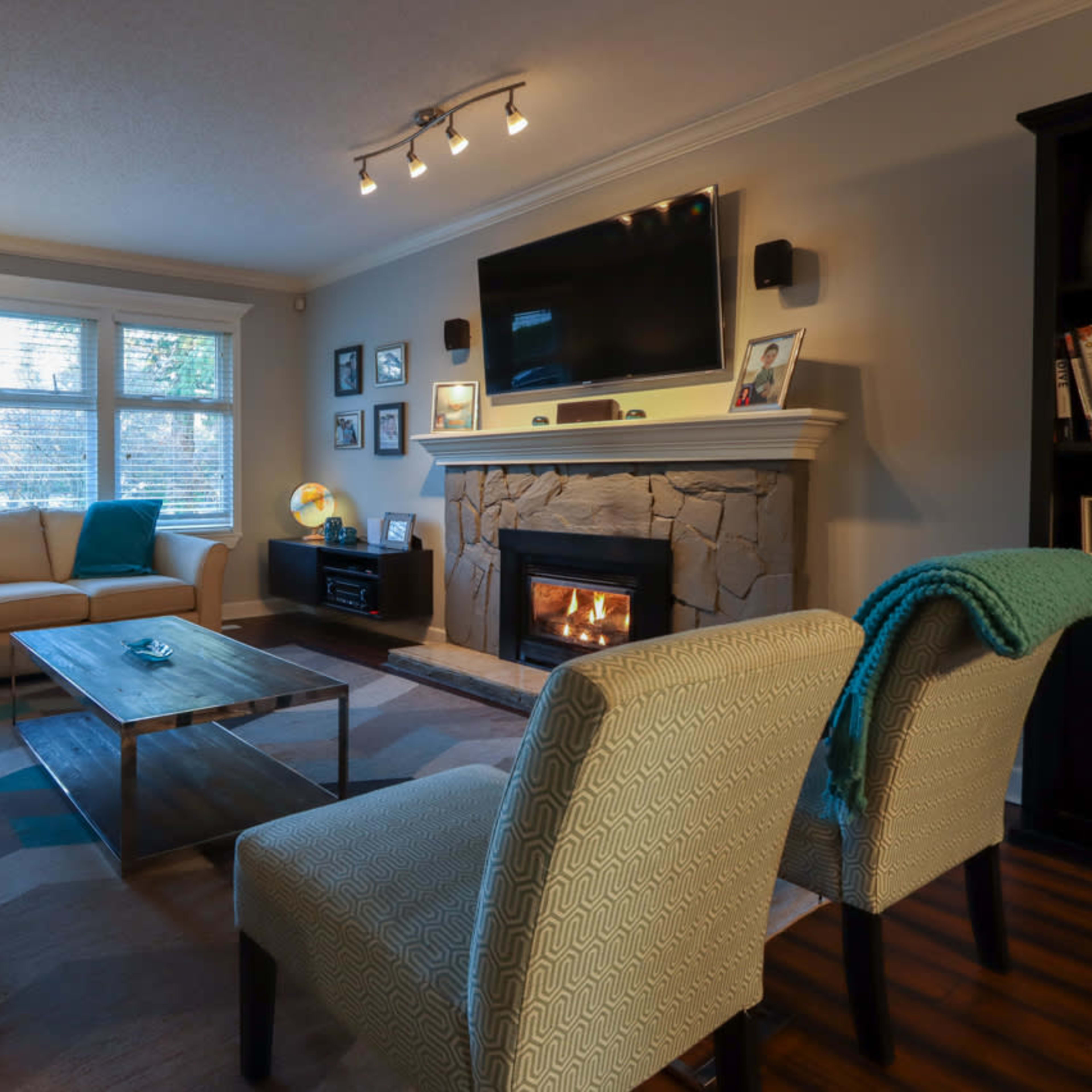 A living room features a beige sofa set, a stone fireplace with a TV mounted above, and a wooden coffee table, illuminated by natural light from the windows.