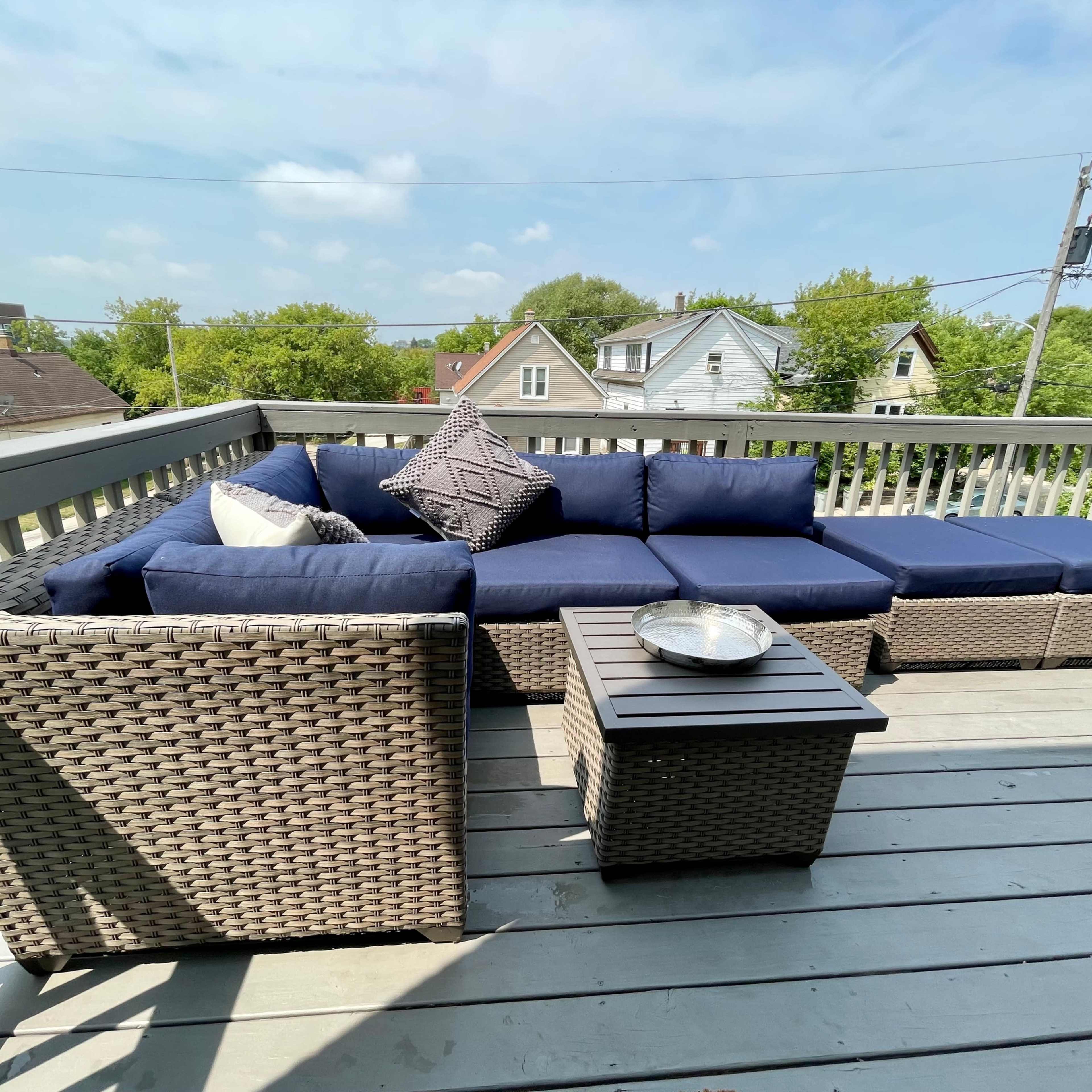 A spacious outdoor deck features a sectional sofa with navy cushions and a small table, surrounded by trees and houses in the background.