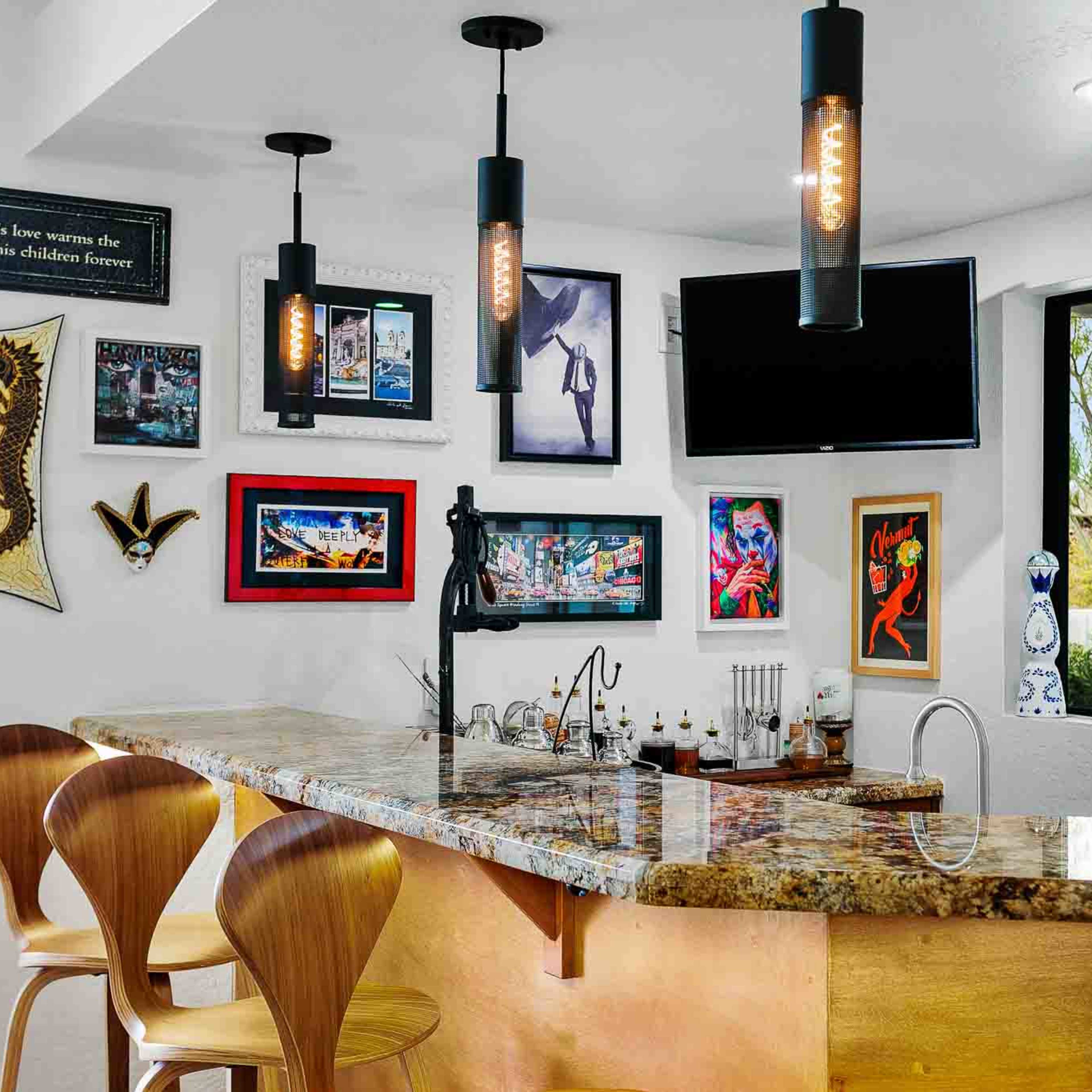 A modern kitchen bar features wooden stools and a granite countertop, surrounded by various framed artworks and a mounted television.