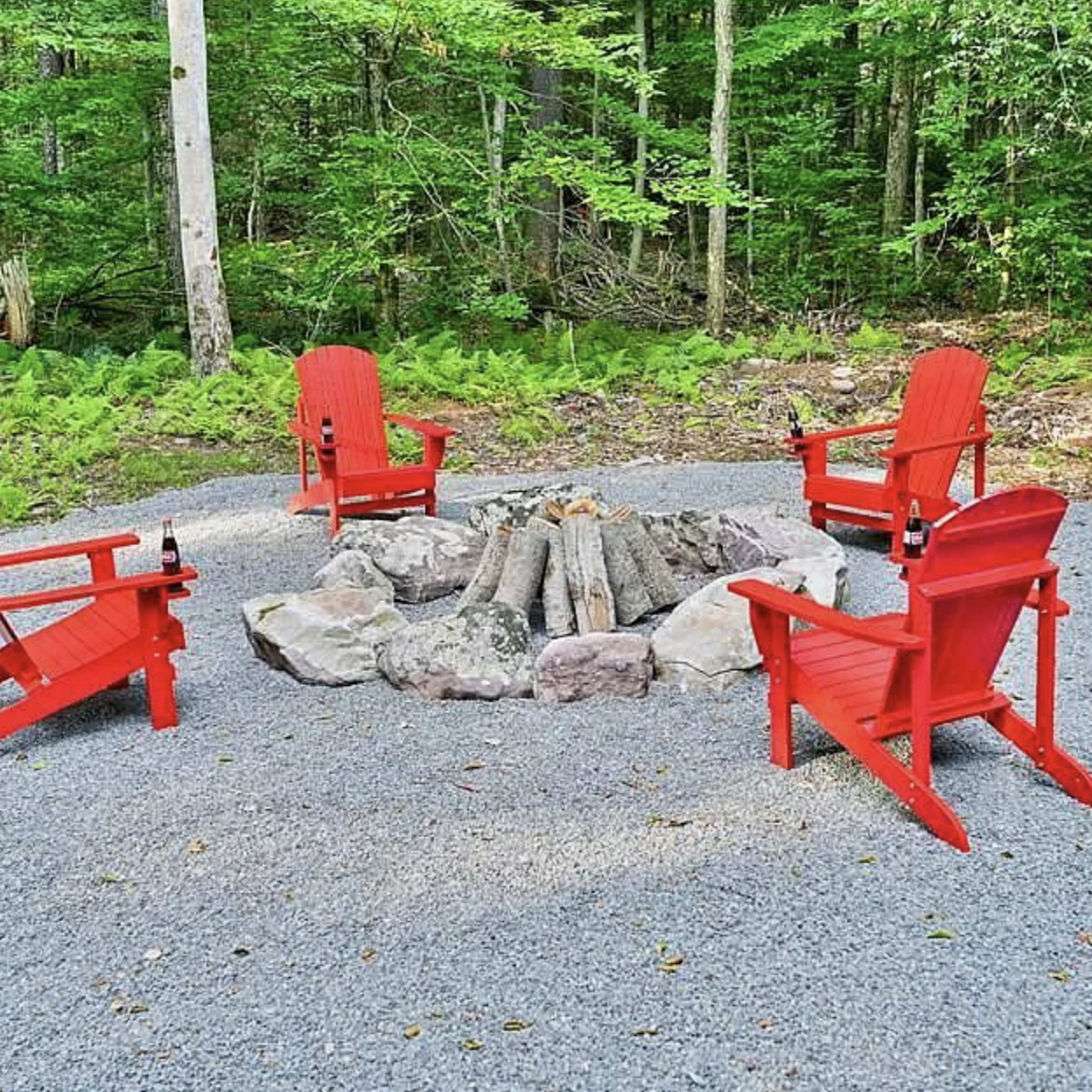 A circular arrangement of four red Adirondack chairs is set around a stone fire pit in a wooded area.