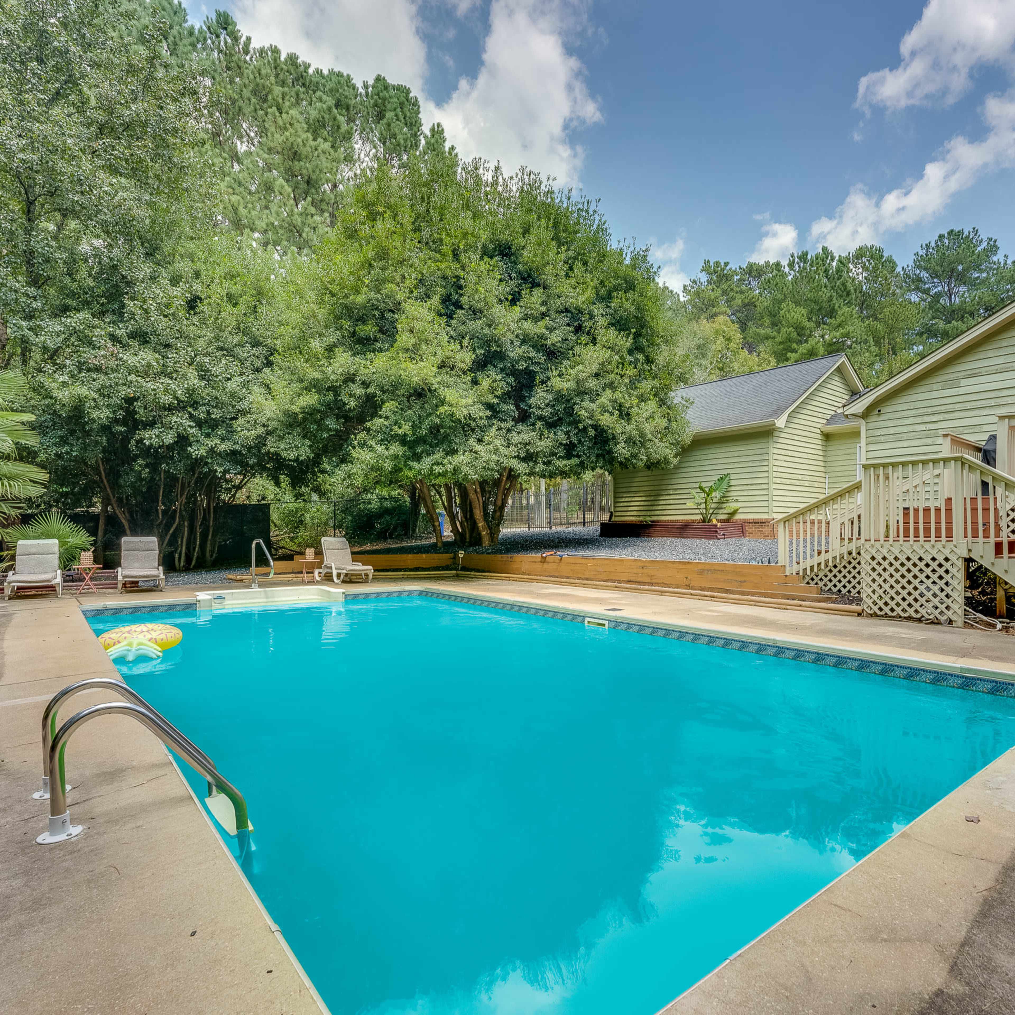 A clear swimming pool surrounded by concrete, with lounge chairs nearby and a house partially visible in the background.
