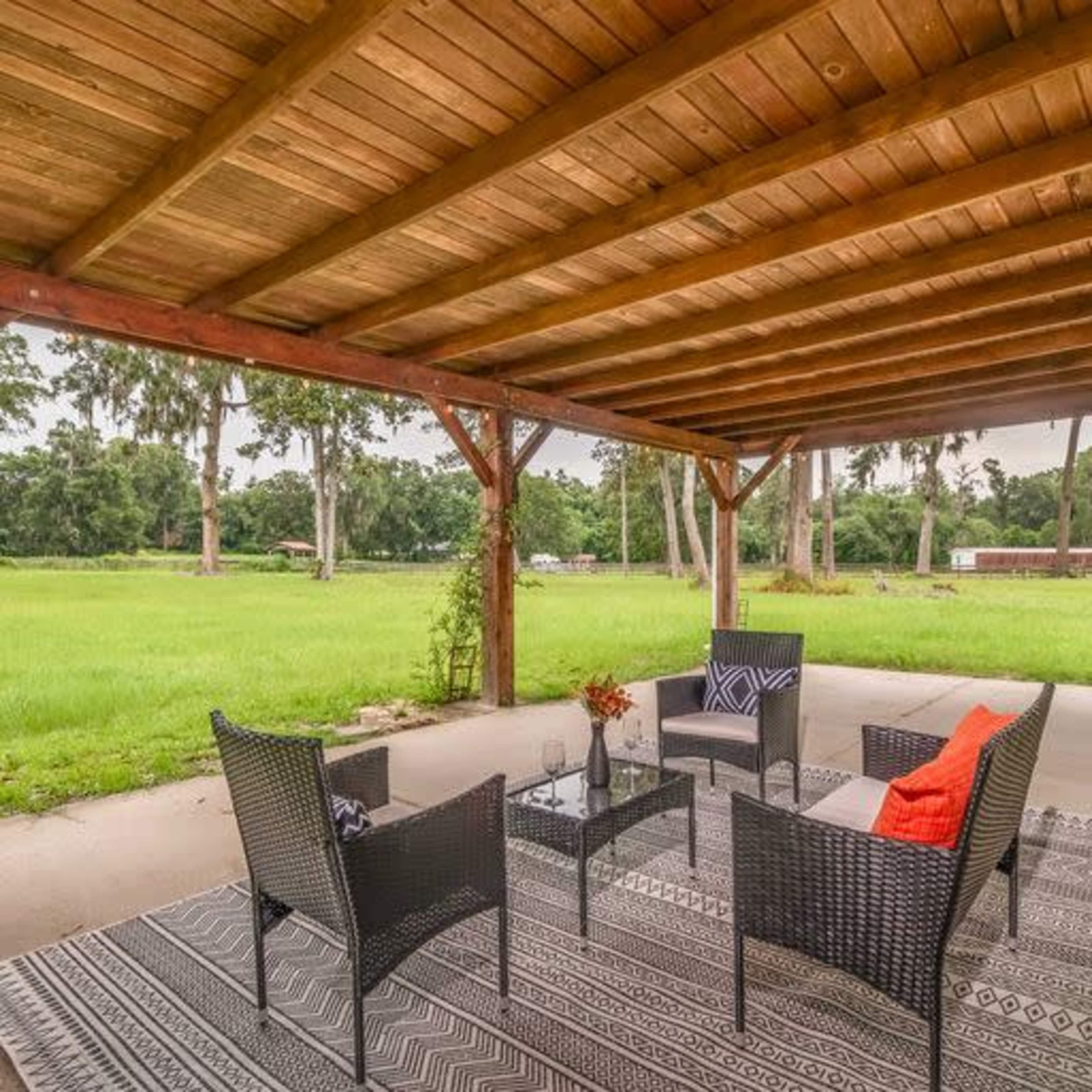 A covered patio features a seating arrangement with four chairs and a table, surrounded by a green landscape and tall trees.