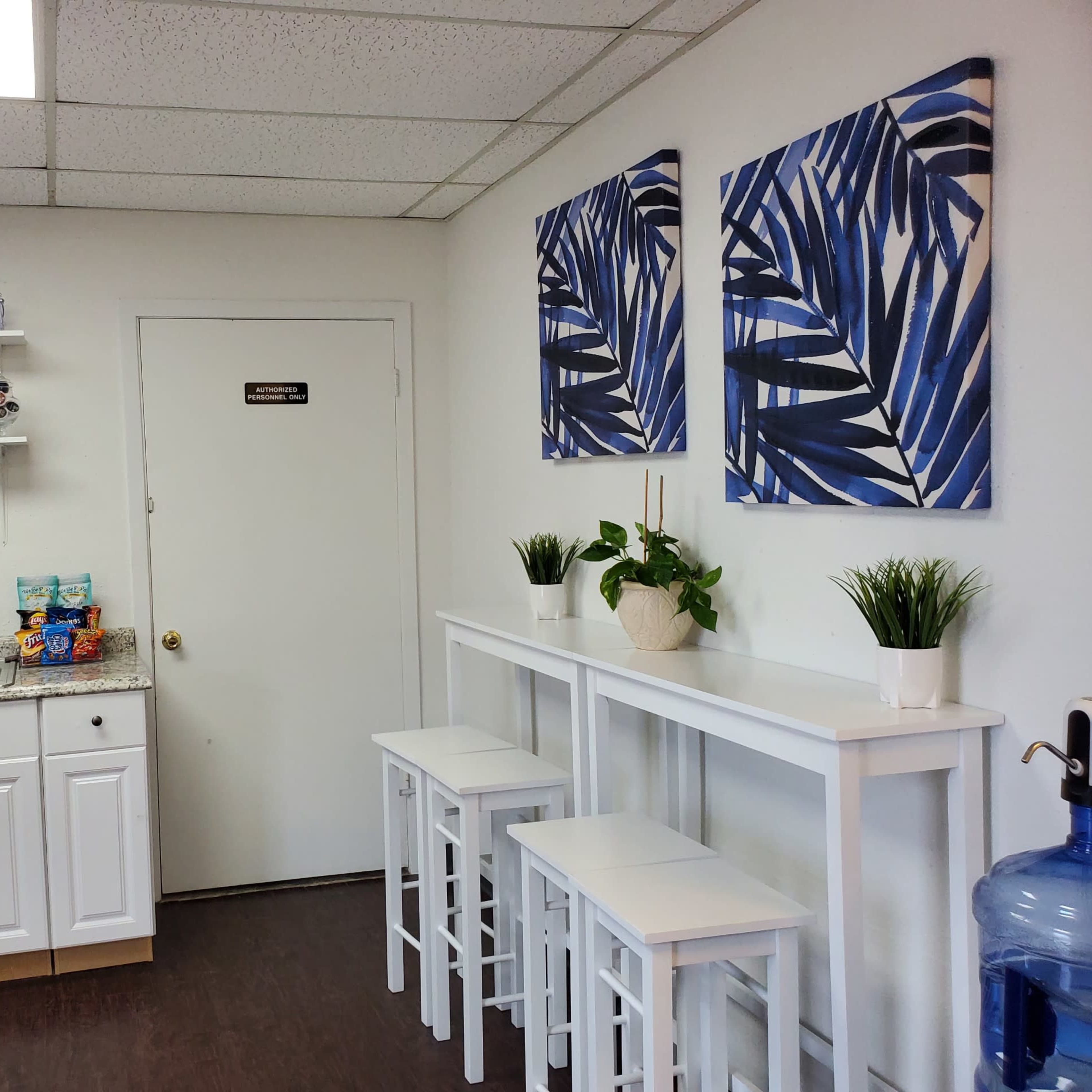 A kitchenette area with a countertop, a sink, two paintings of blue leaves on the wall, a small table with three stools, and a water dispenser.