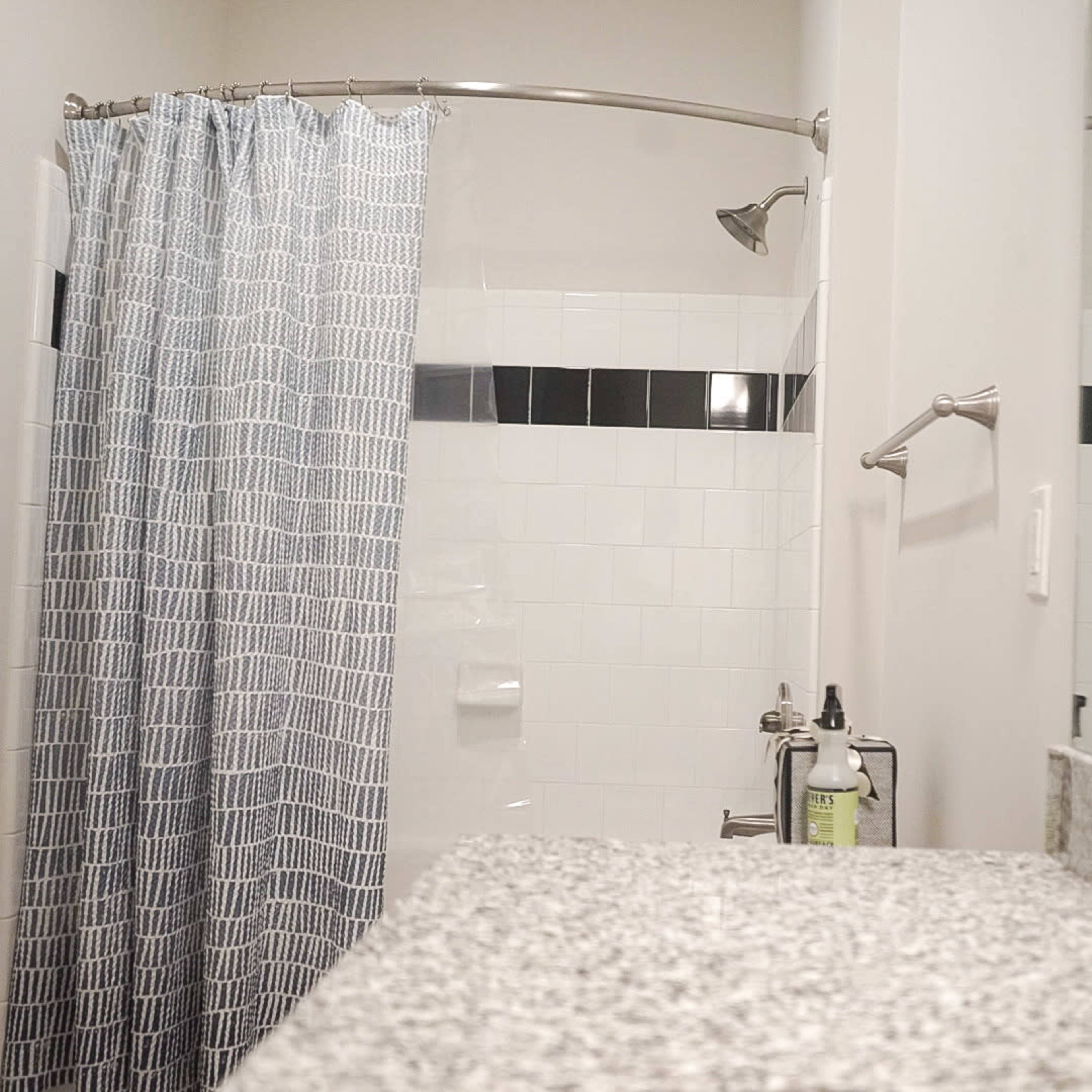 A bathroom features a shower with a curved curtain rod, gray and white patterned shower curtain, and a countertop with toiletries.