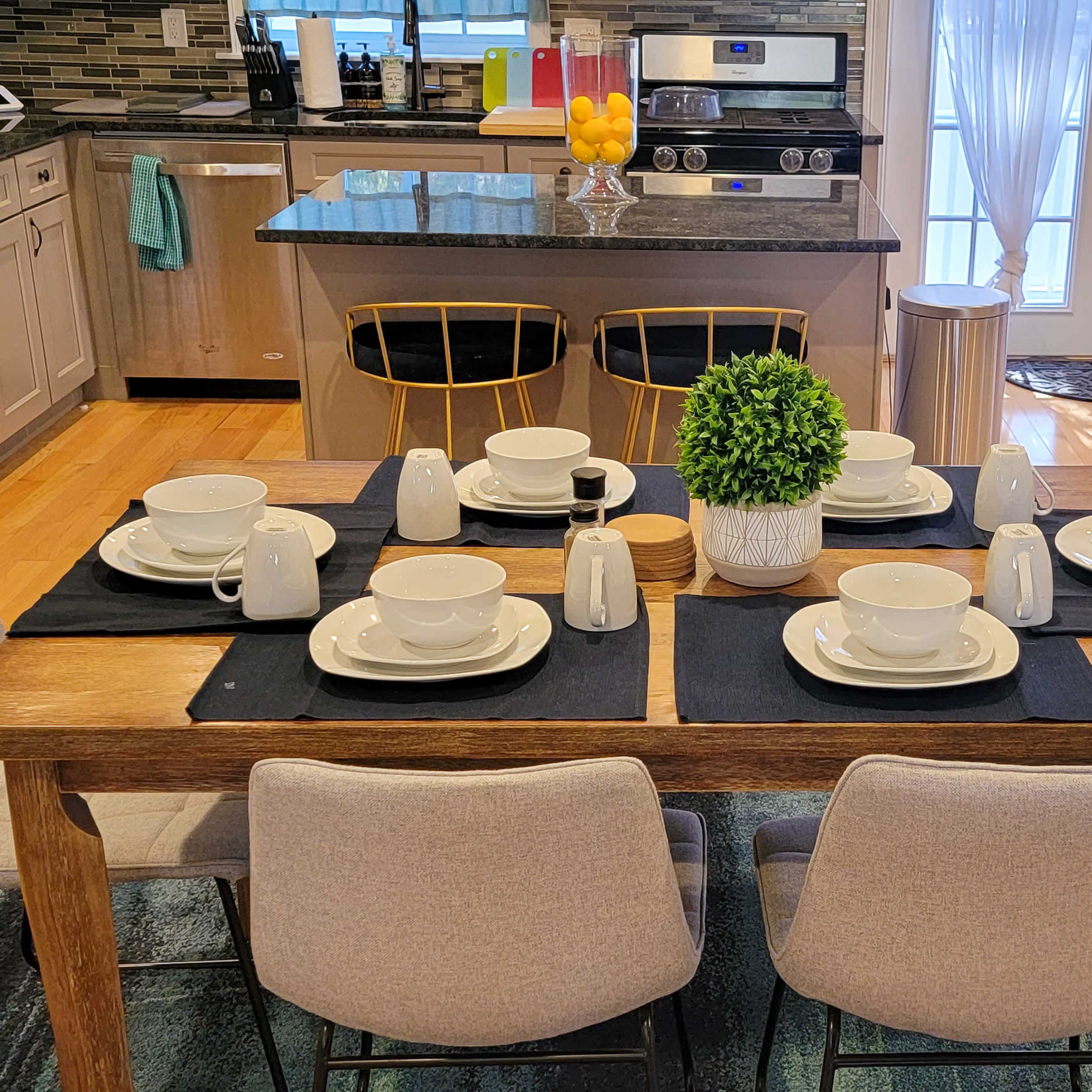 A dining table is set for six with plates, cups, and a small plant centerpiece, situated in a kitchen that features modern appliances and a breakfast bar.