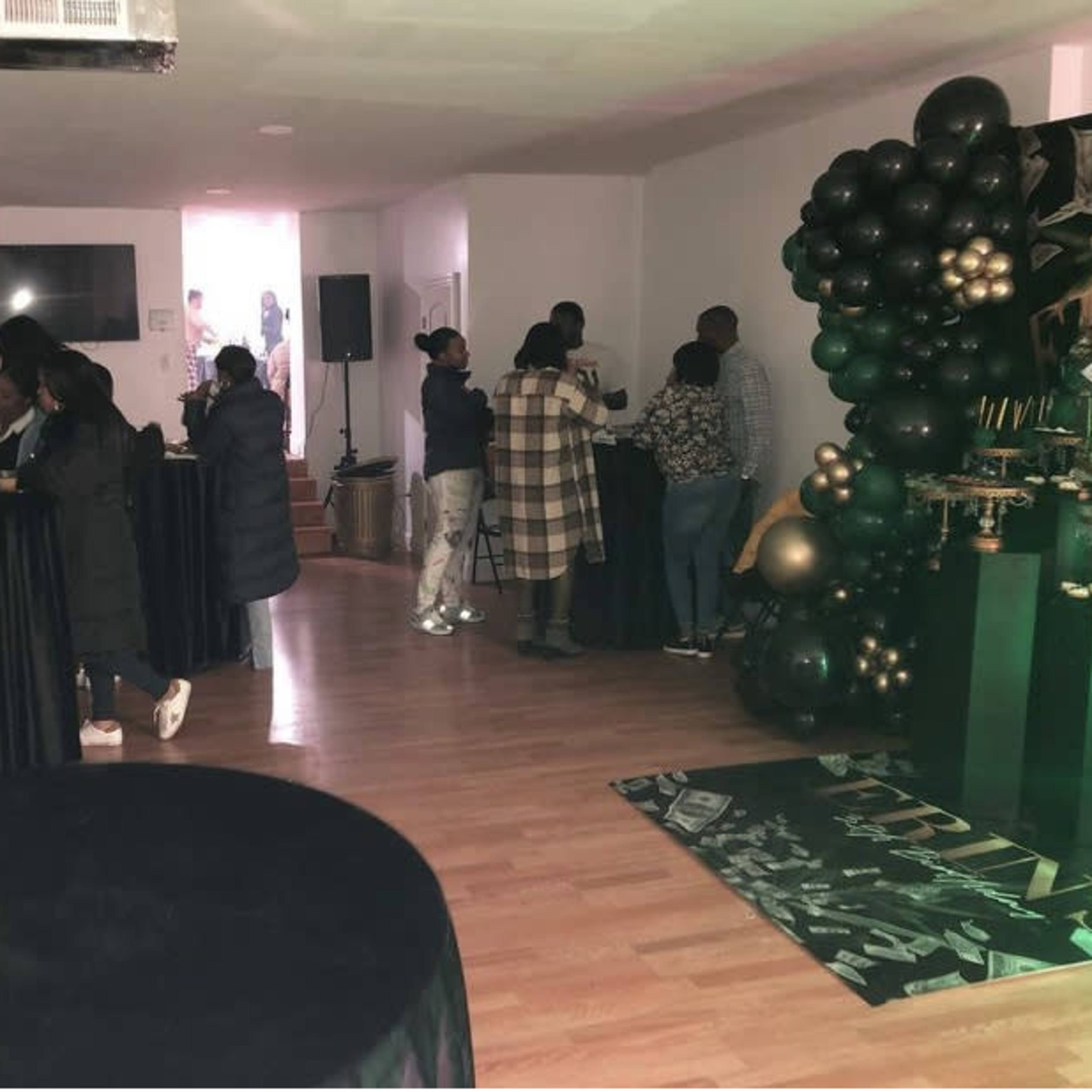 A gathering in a modern indoor space with guests socializing at black tables and a decorative balloon display in the corner.
