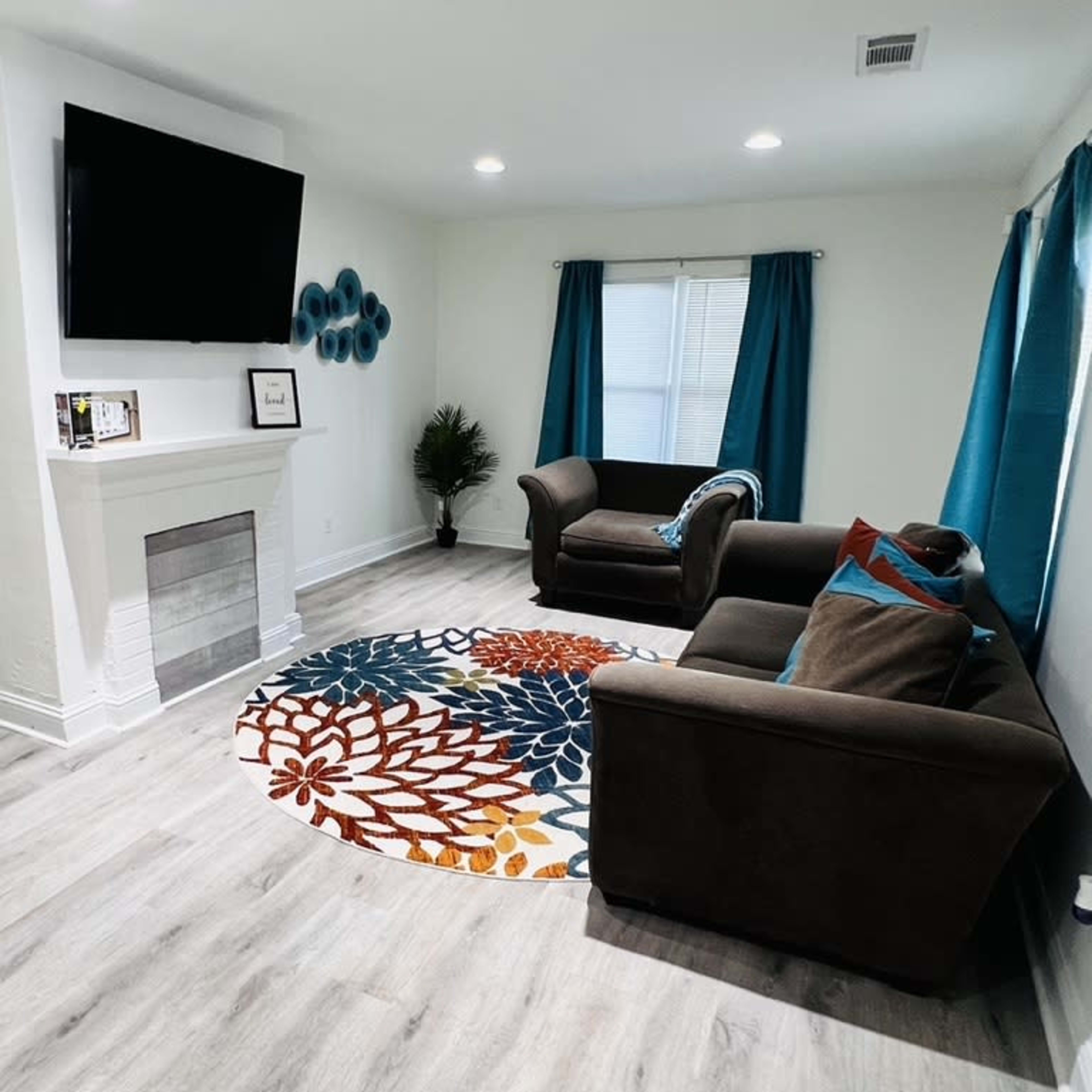 A modern living room with two brown sofas, a colorful circular rug, a television mounted on the wall, and blue curtains covering the windows.