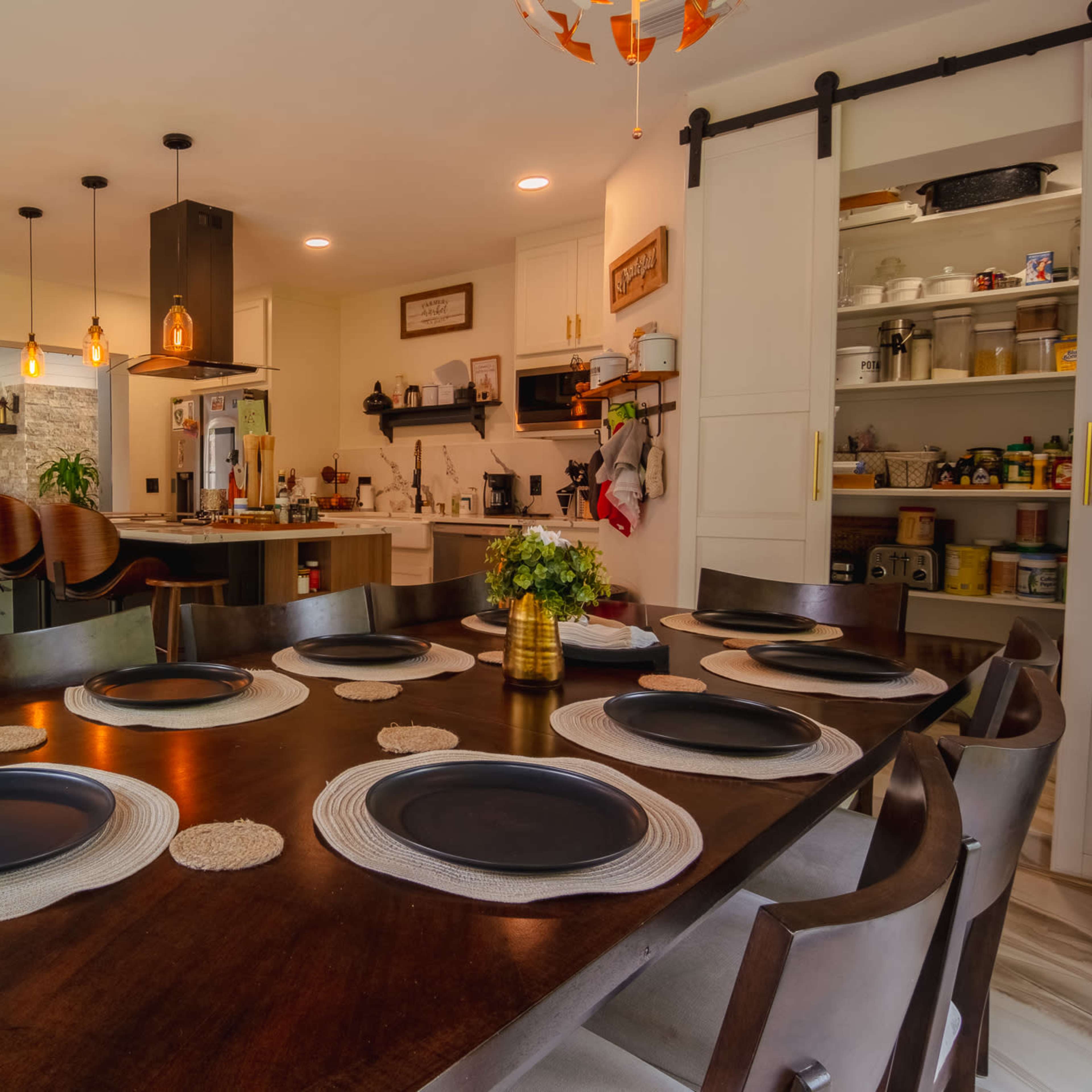 A dining table with black plates and woven placemats is set in a modern kitchen and dining area featuring pendant lighting and an open layout.