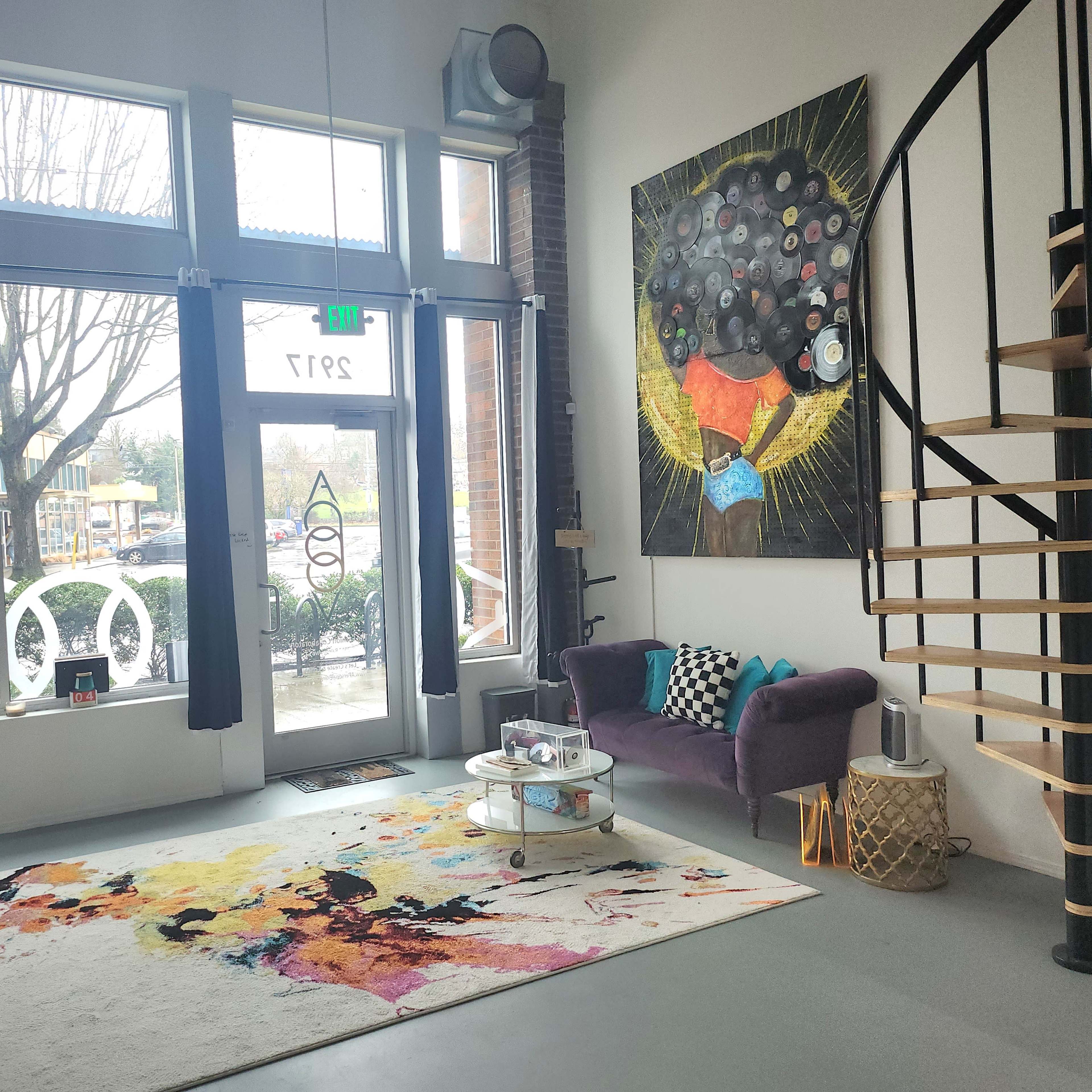 A spacious, modern room with large windows, a purple sofa, a colorful area rug, and a staircase leading to an upper level, accompanied by a vibrant wall art piece.