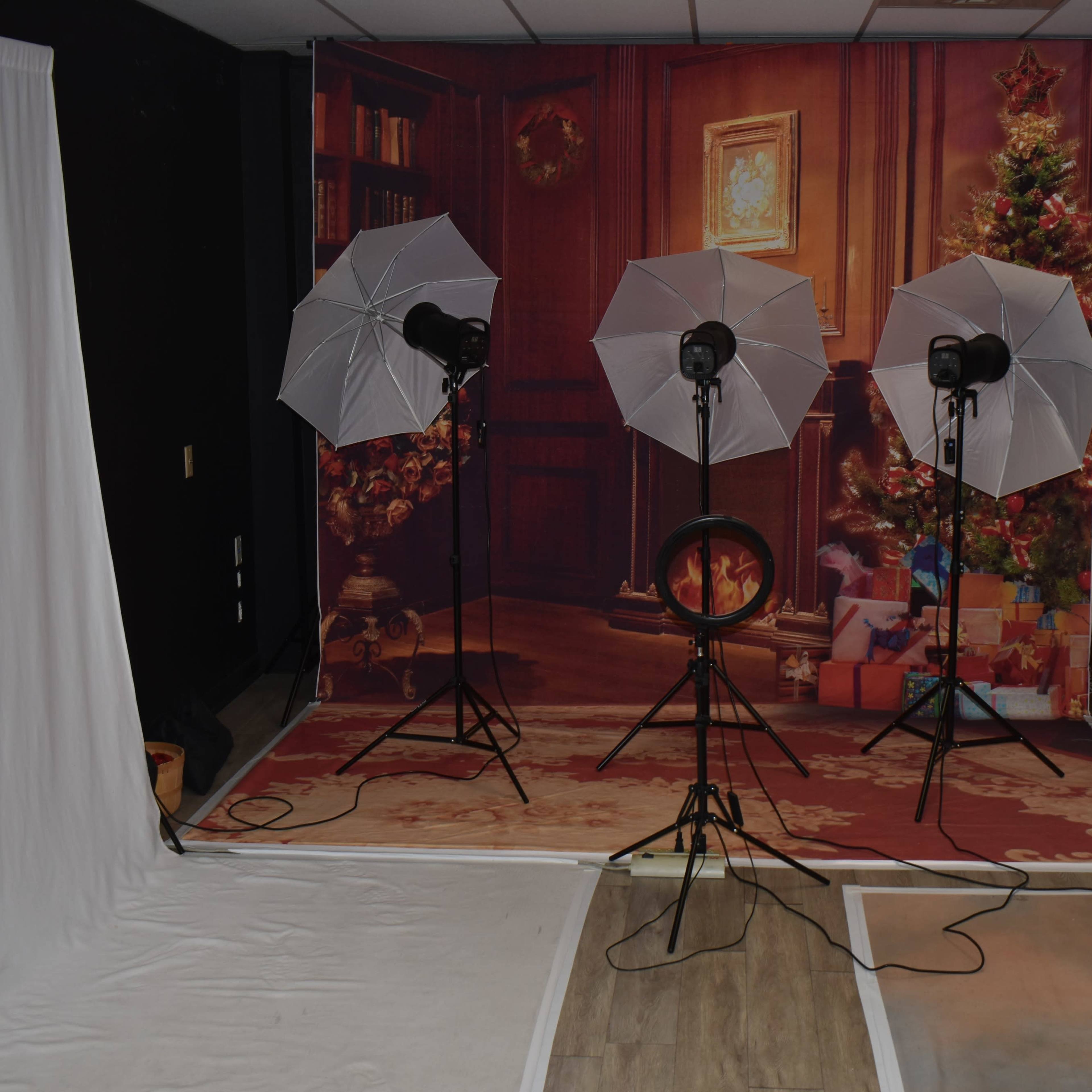 A photography studio setup with three lights on tripods, a backdrop featuring a decorated room with a Christmas tree, and various fabric drapes on the floor.