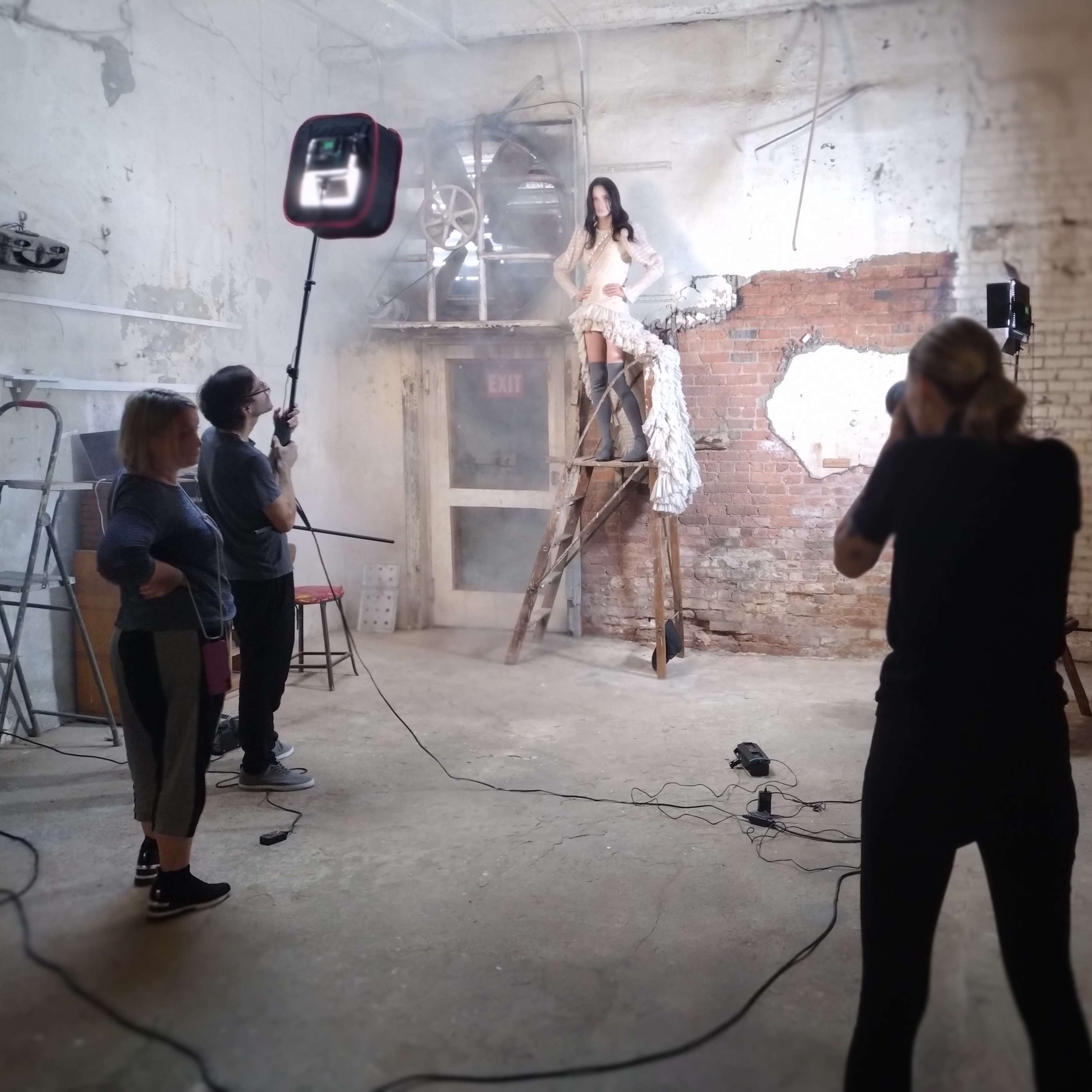 A model sits atop a ladder in an industrial space while a photographer captures the shot and crew members adjust lighting equipment.