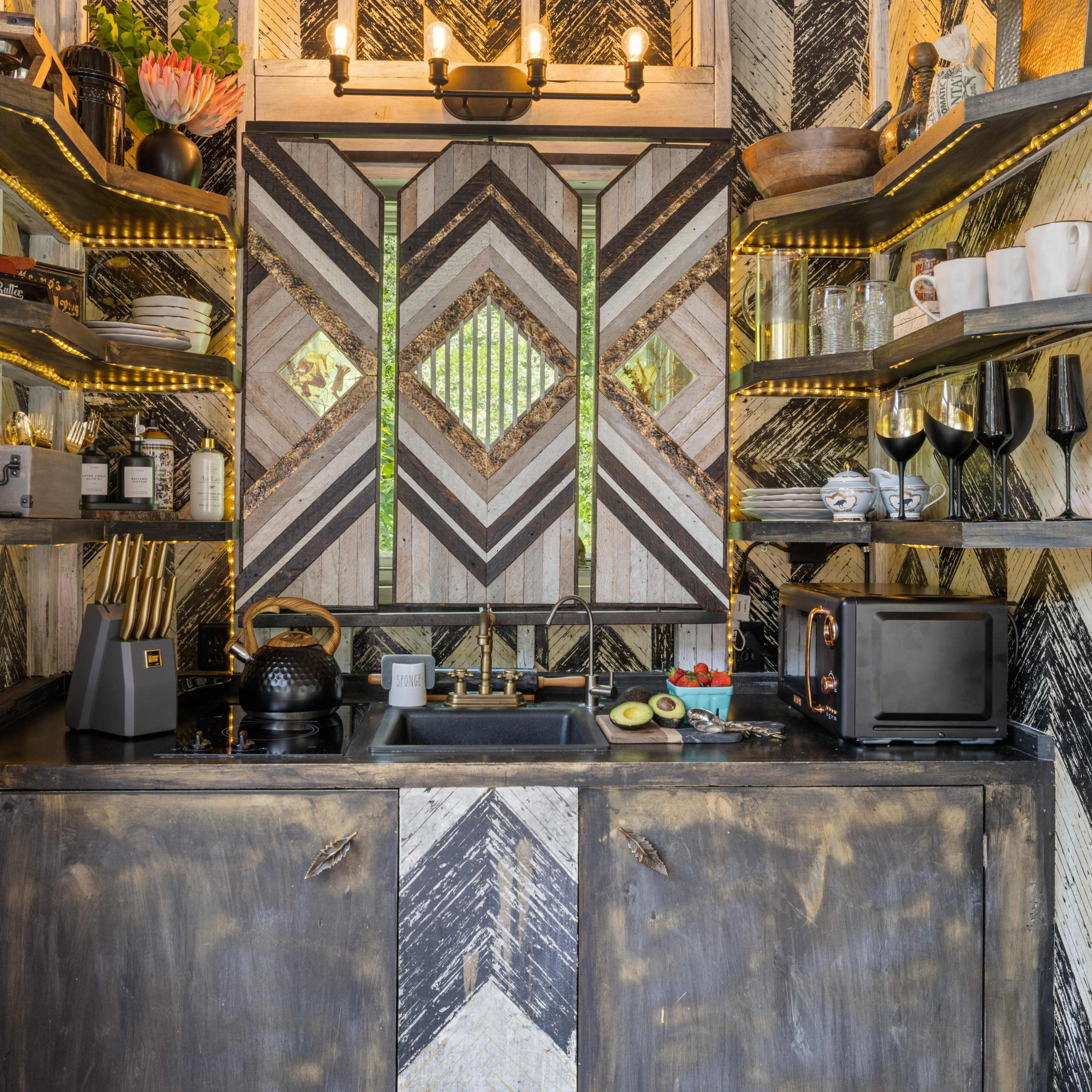 A compact kitchen area featuring a dark wood cabinetry with geometric-patterned walls, shelves holding various dishes and decor, and a small sink with appliances and fresh produce on the counter.