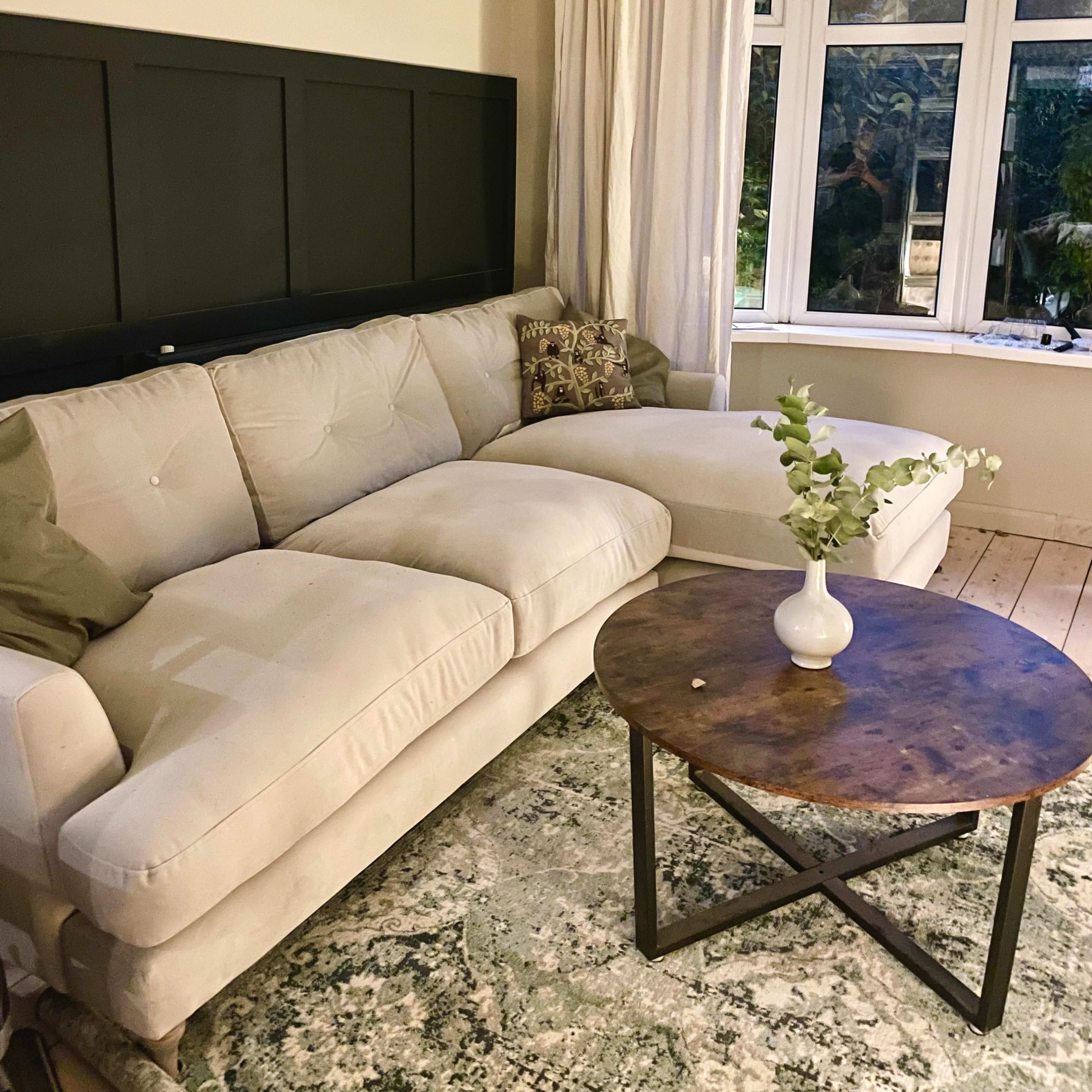 A light-colored sectional sofa with a decorative cushion is positioned next to a round wooden coffee table in a room with a large window and sheer curtains.