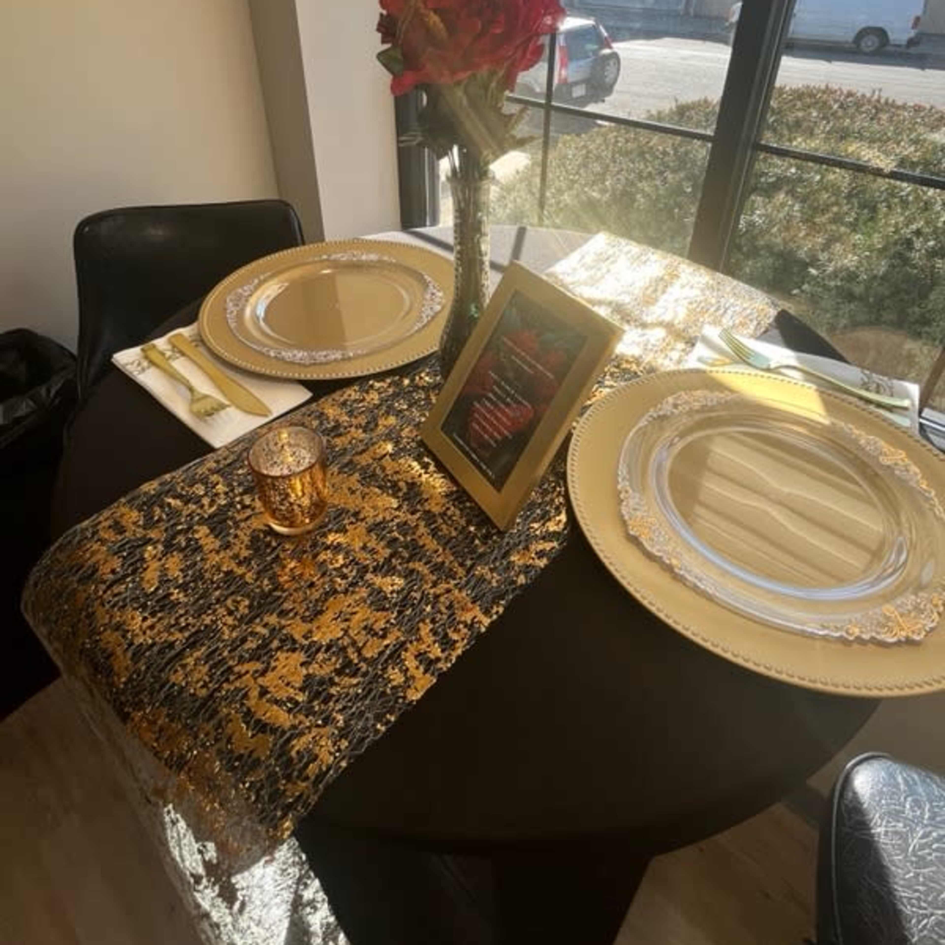 A elegantly set dining table with two plates, a vase of red flowers, a framed menu, and a decorative runner under natural light.