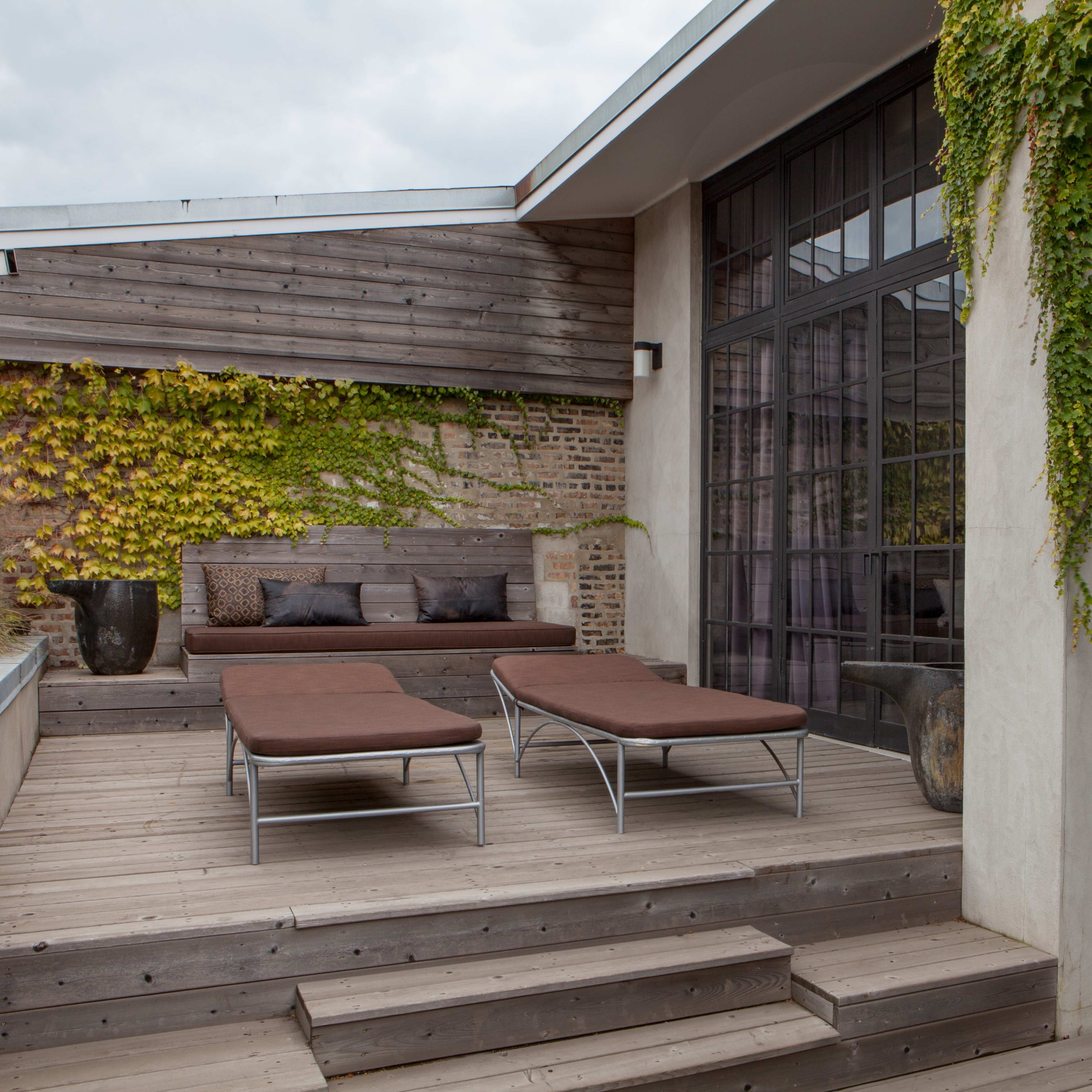 A modern outdoor terrace with two brown lounge chairs, wooden decking, and a backdrop of green climbing vines on the building.