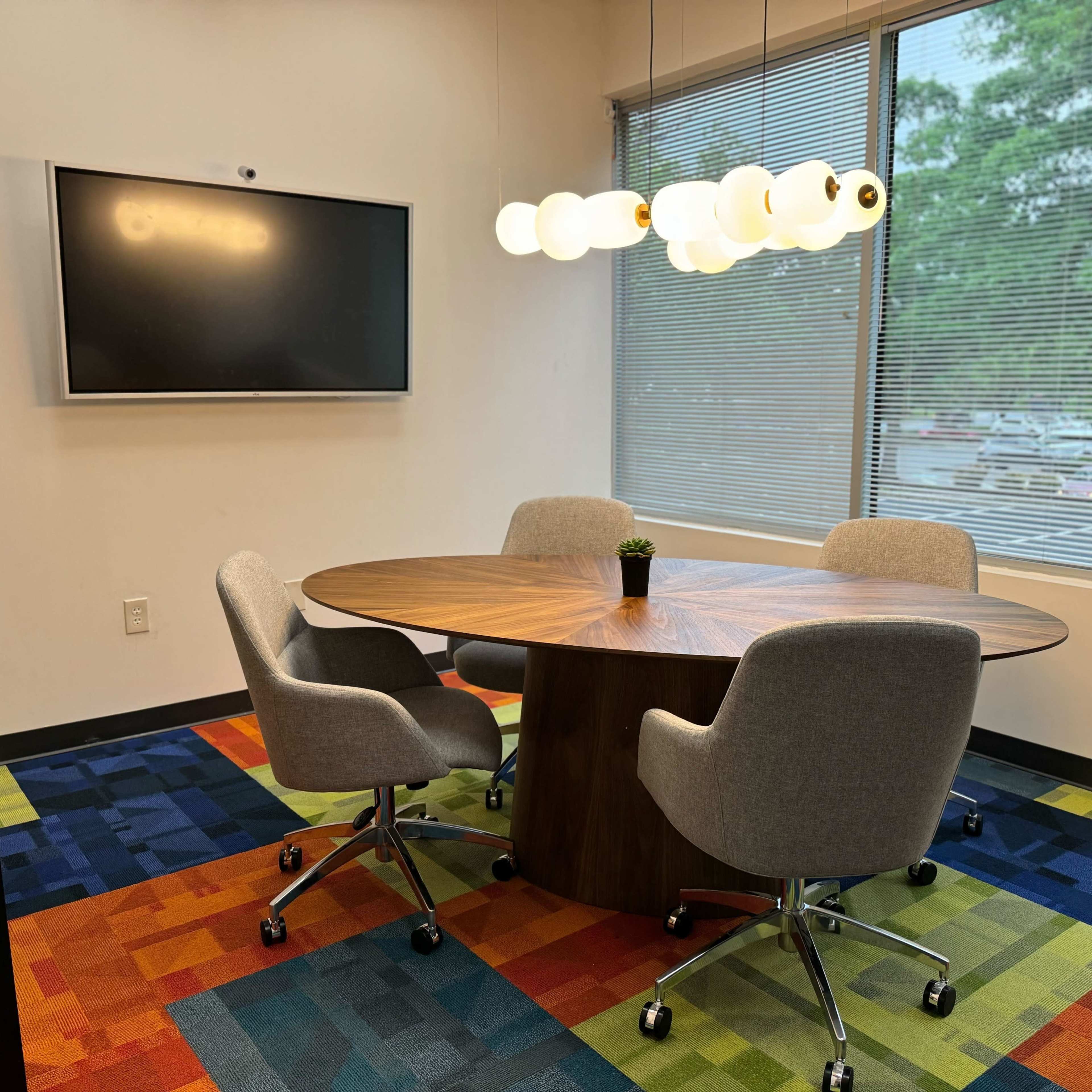 A modern conference room features a round wooden table surrounded by four gray chairs, with a large screen on the wall and a colorful carpet.