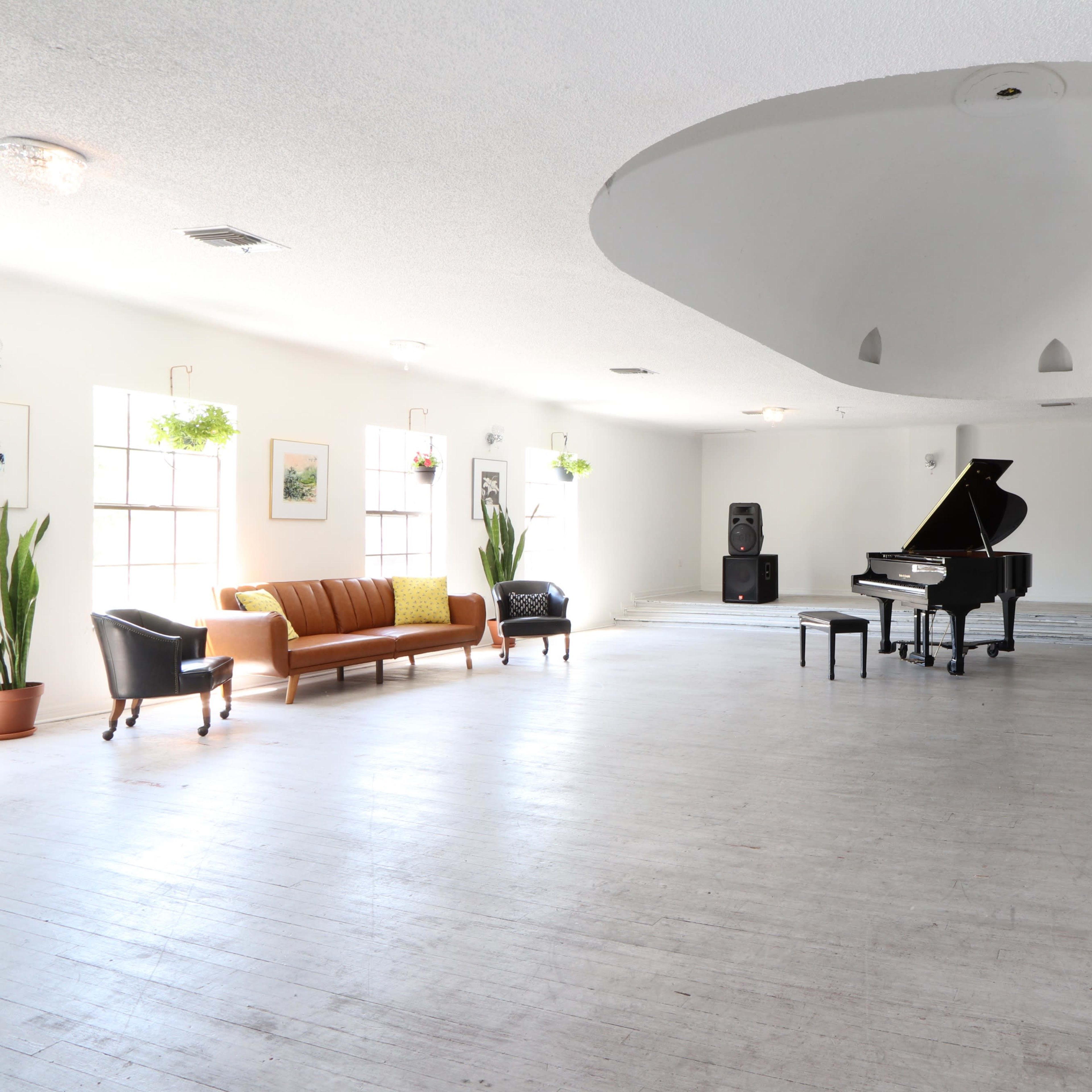 A spacious, empty room features a grand piano surrounded by a sofa, a chair, and two large speakers, with light streaming in through several windows.