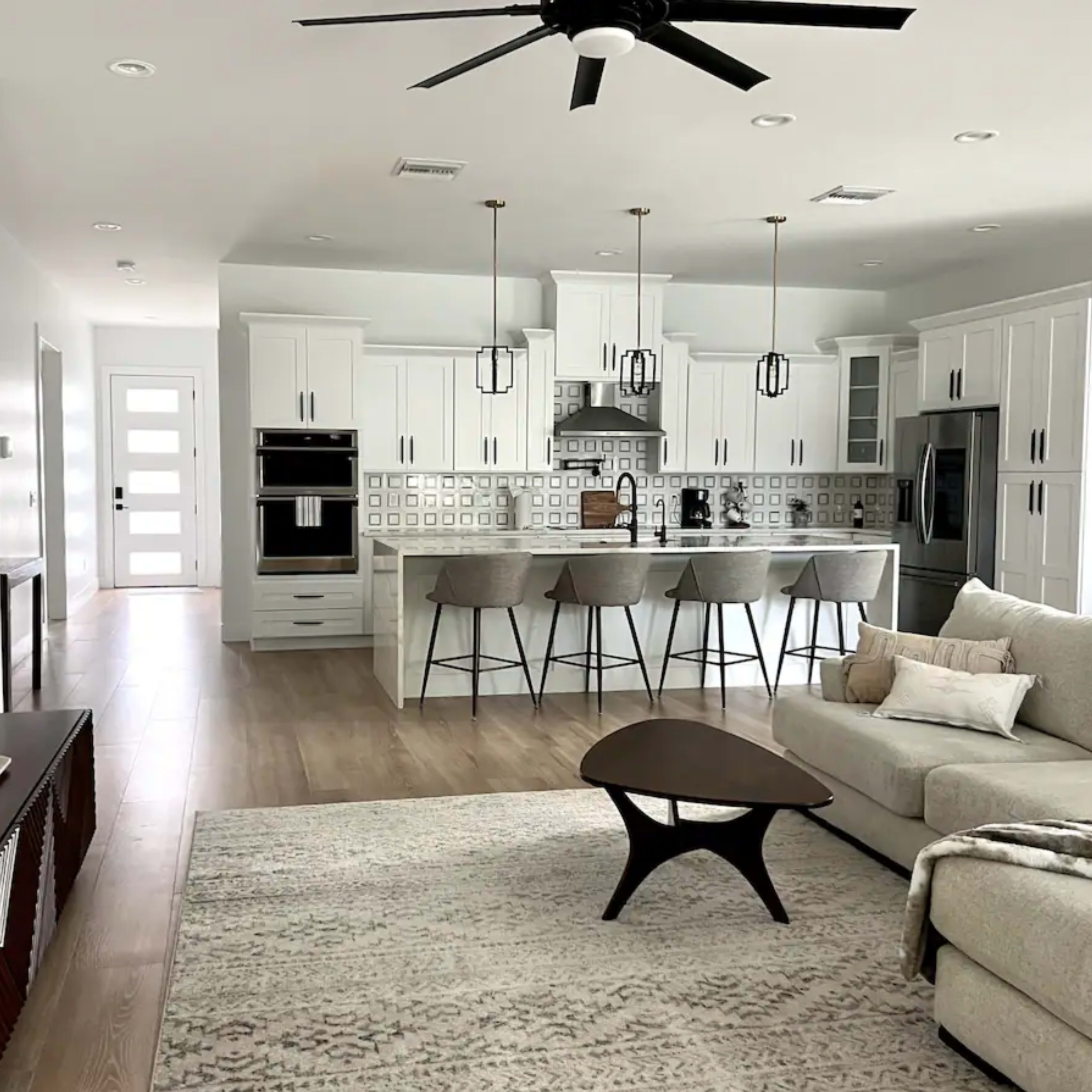 A modern living room features a light-colored sofa, a coffee table, and a view of a kitchen with white cabinets and pendant lights.