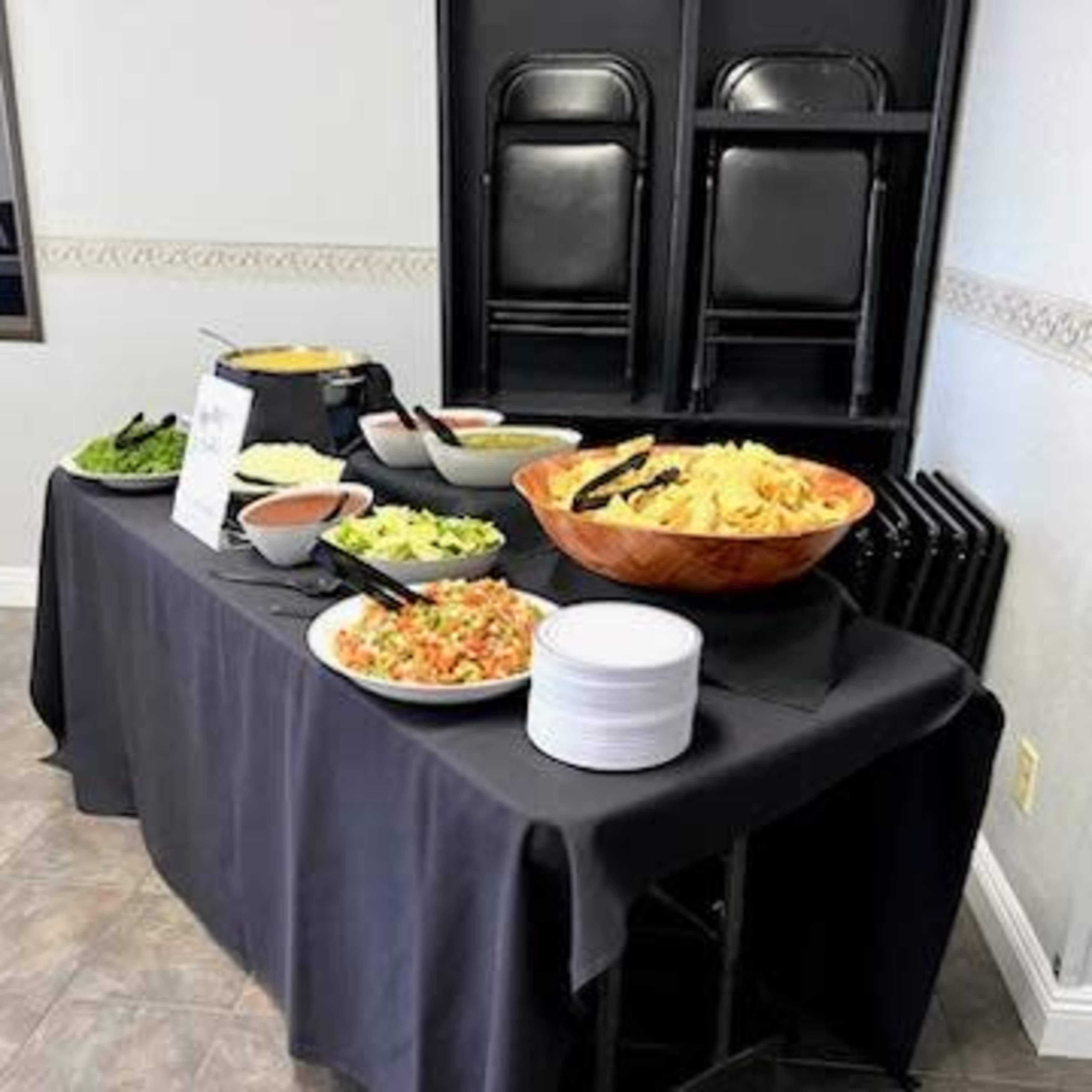 A buffet table is set up with various bowls of salad, chips, and sauces, alongside a stack of white plates, in a room with folding chairs leaning against the wall.