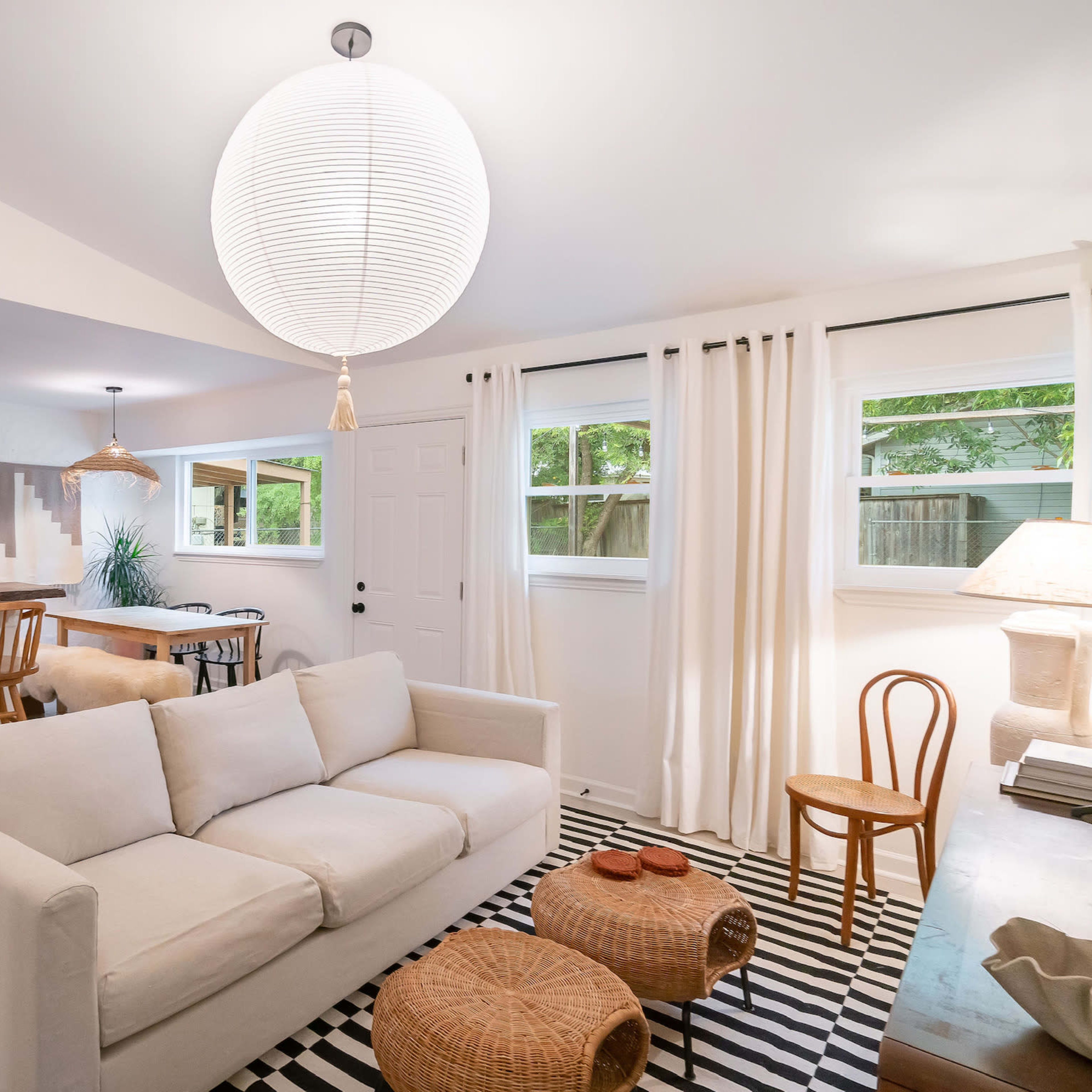 A modern living area with a white sofa, a black and white striped rug, a dining bar with wooden stools, and large windows allowing natural light.