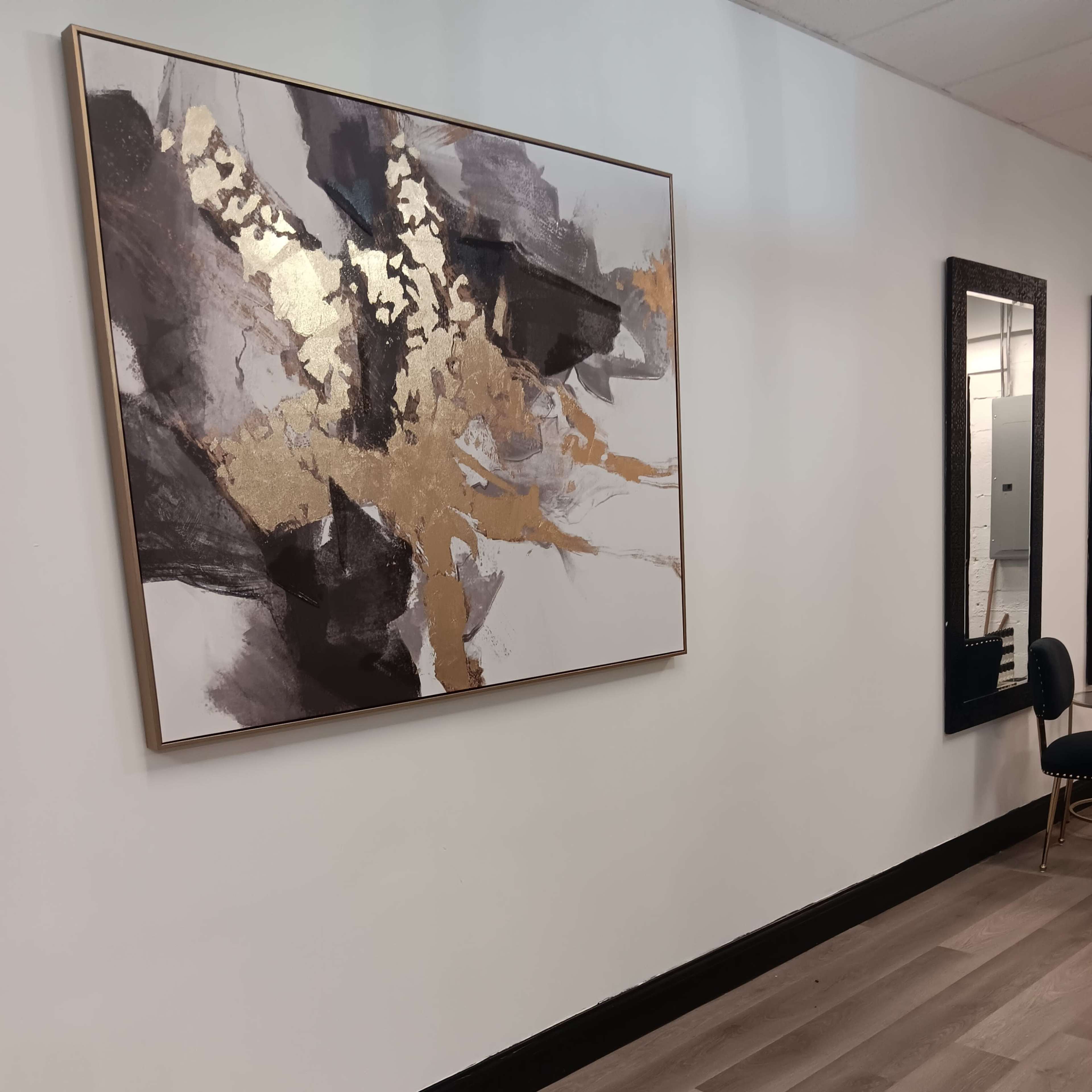 A spacious interior with a large abstract painting featuring gold accents on a white wall, alongside a tall mirror and a black chair on a wooden floor.