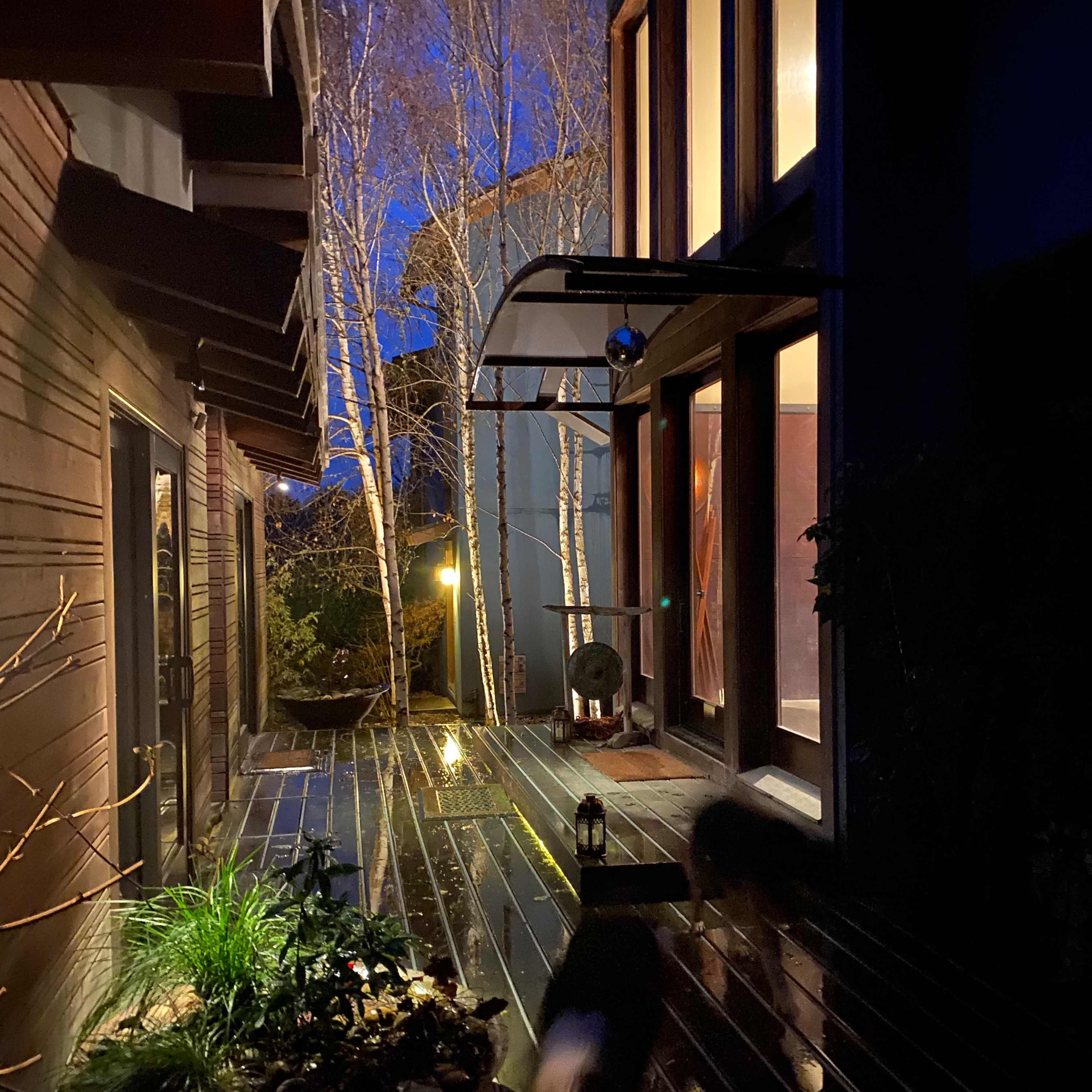 A pathway with wooden decking runs between two buildings, leading to a well-lit entrance while birch trees are visible in the background under a twilight sky.