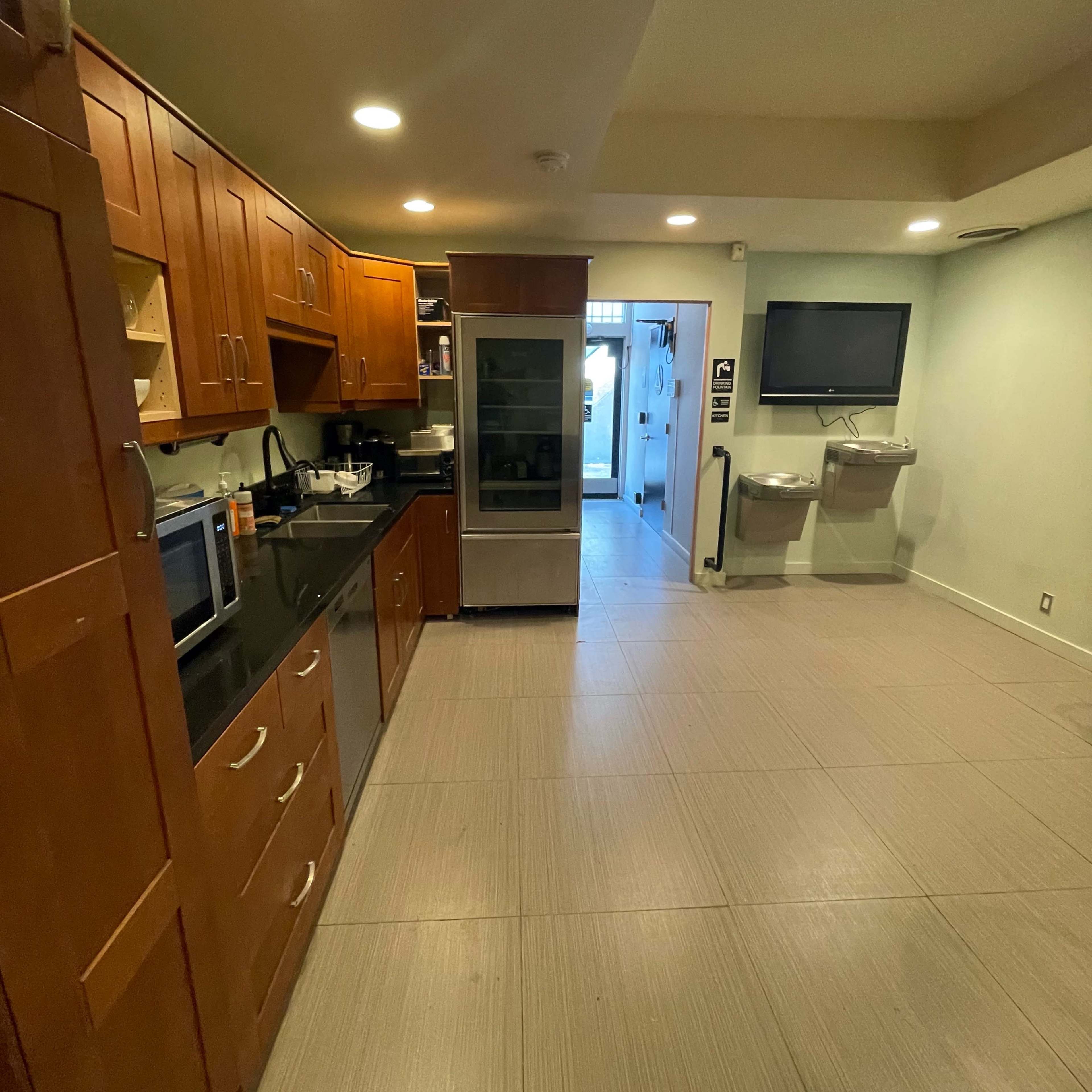 A modern kitchen area with wooden cabinets, a black countertop, a refrigerator, a microwave, and a door leading to an outdoor space, alongside two water fountains.