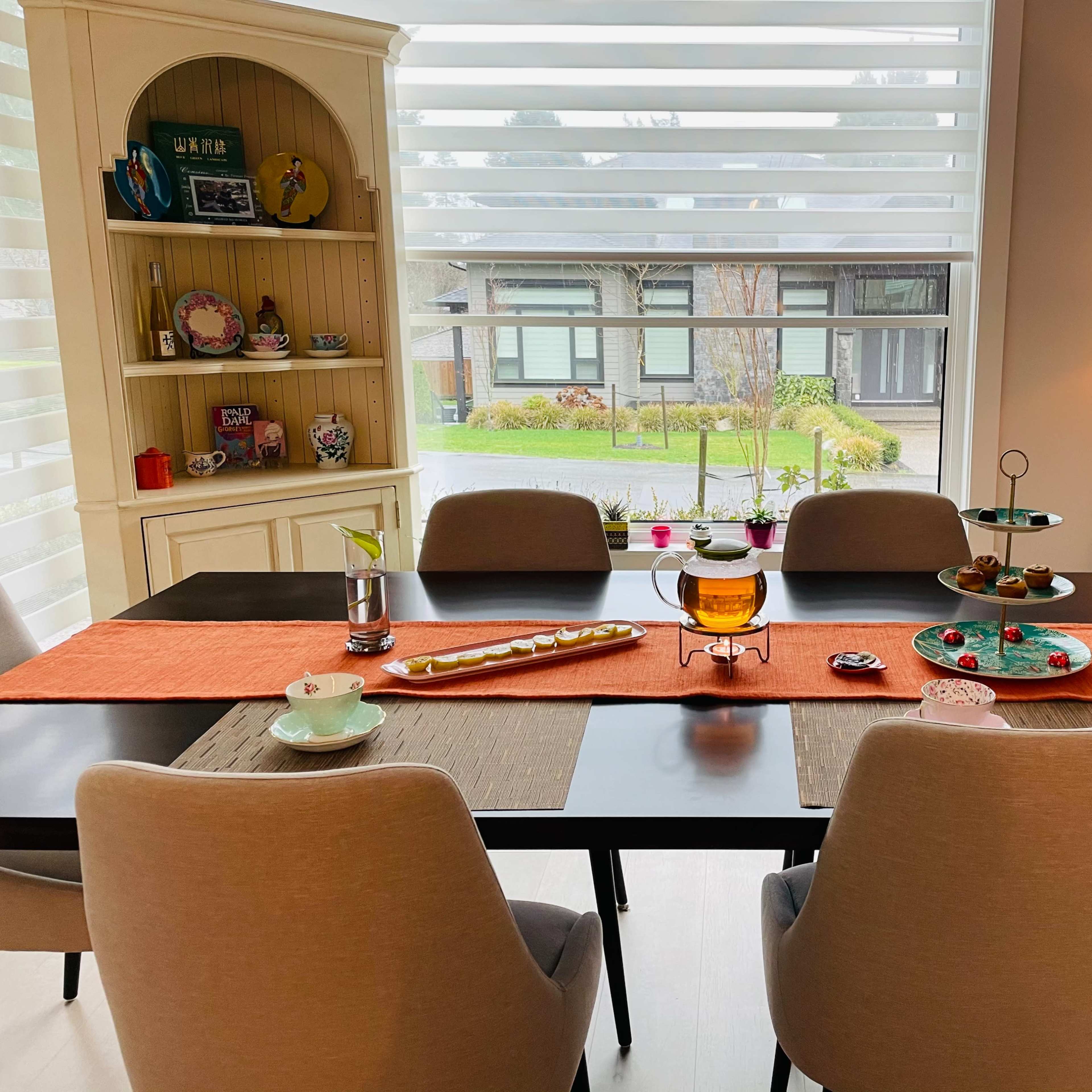 A dining table is set with various items, including a centerpiece and table runners, surrounded by chairs in a brightly lit room with large windows.