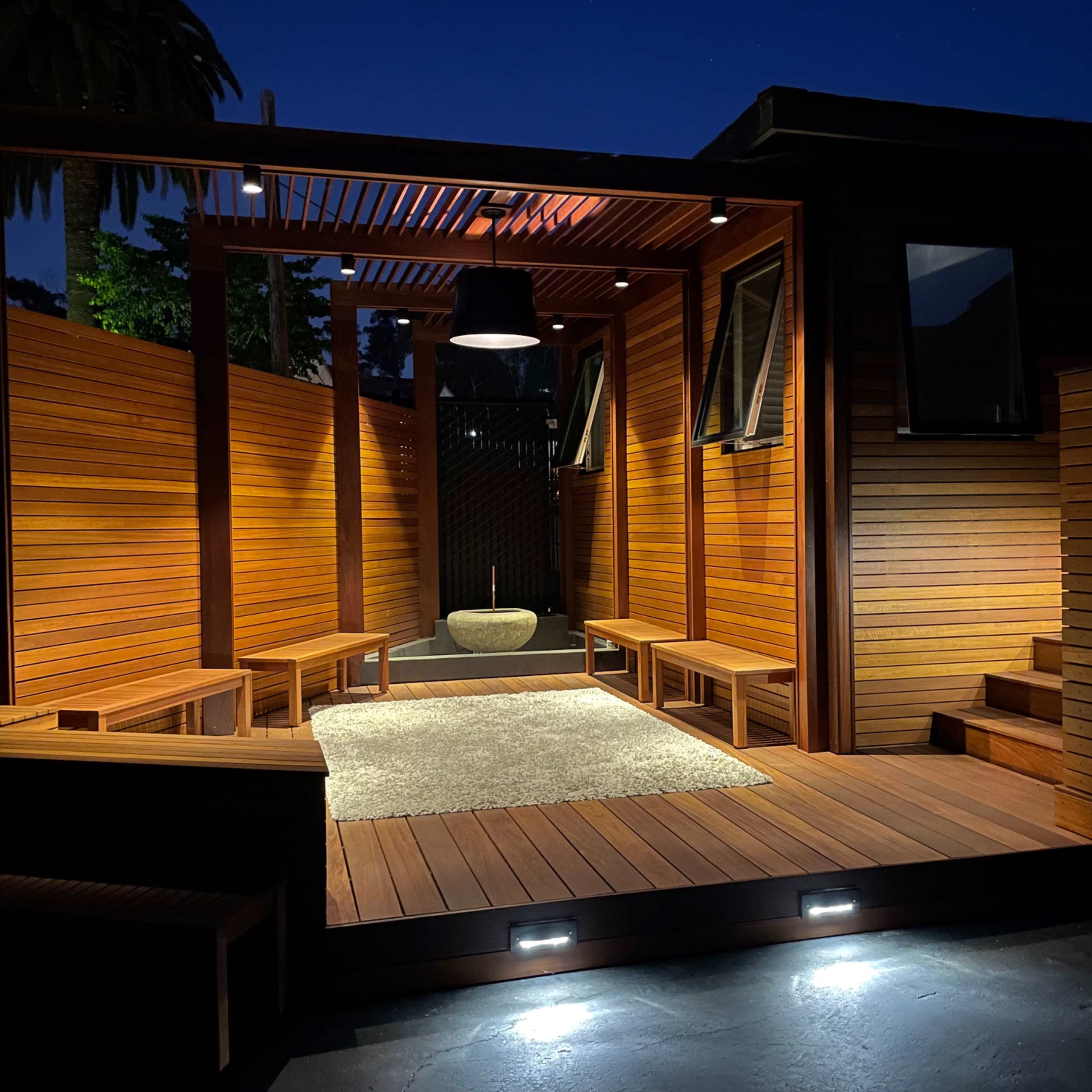 A wooden patio area illuminated at night, featuring a central stone basin, seating arrangements, and wall-mounted windows.