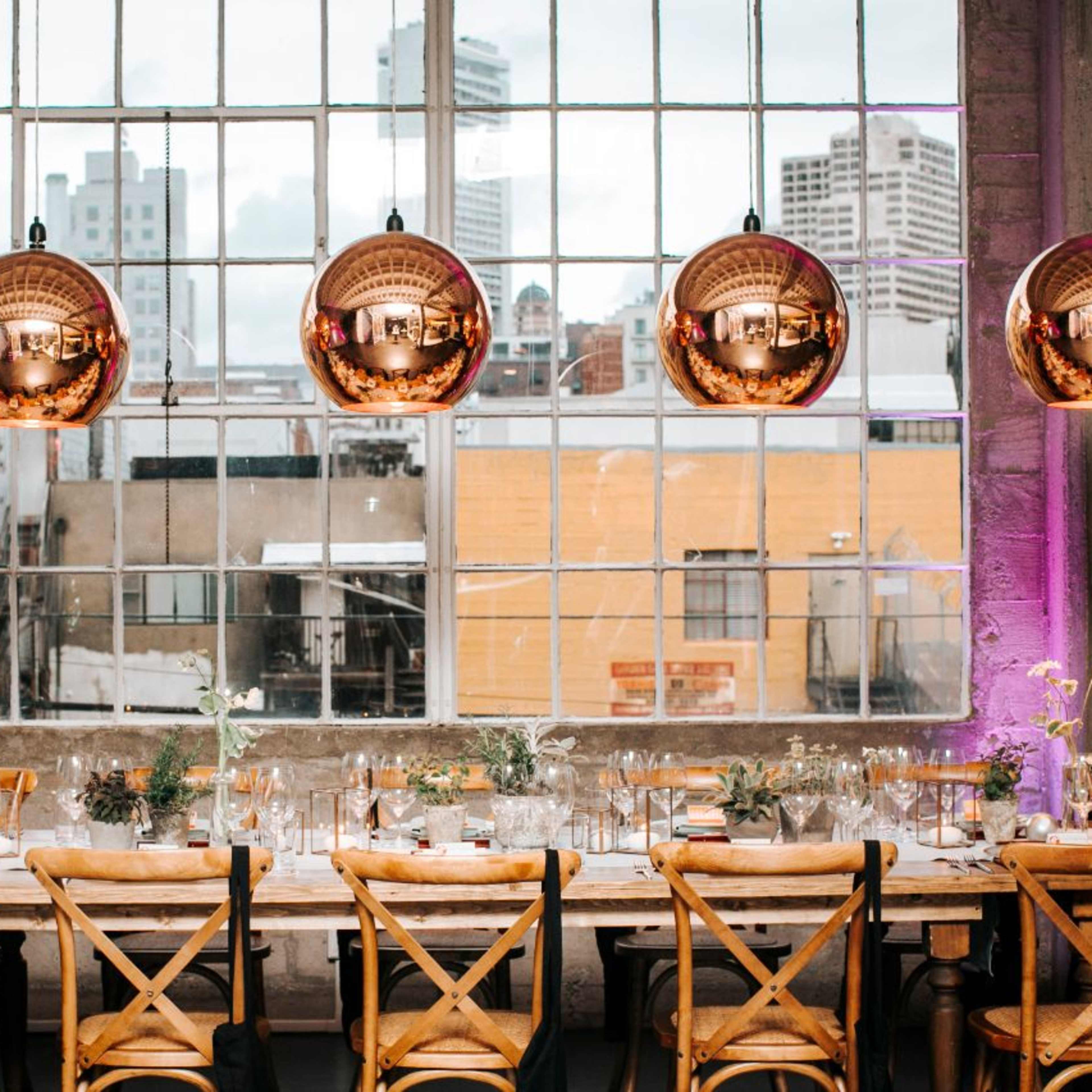A long dining table is set with glasses and flowers, illuminated by large, round pendant lights hanging from the ceiling near a large window overlooking a cityscape.