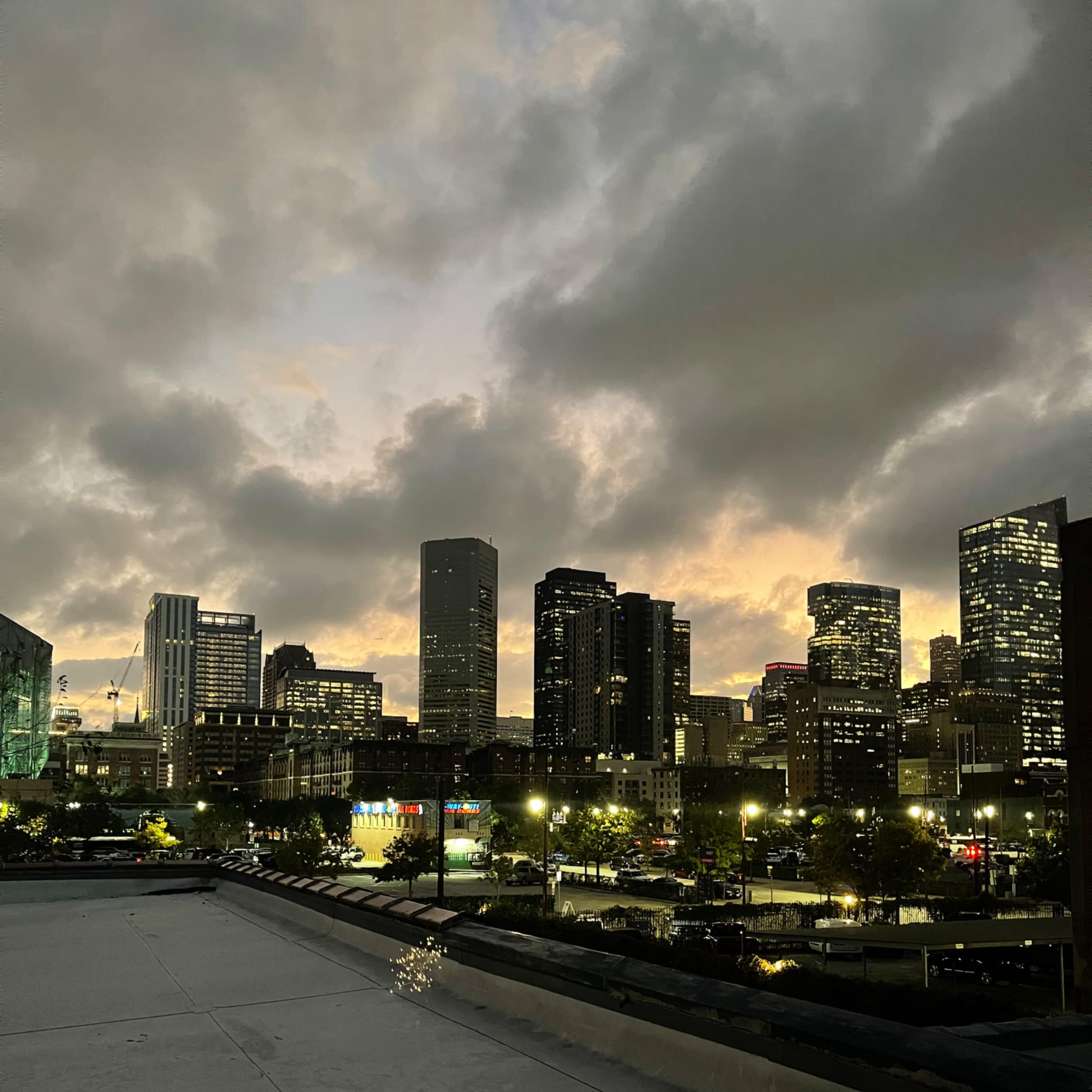 The view shows a city skyline at dusk, with tall buildings silhouetted against a cloudy sky and streetlights illuminating the foreground.