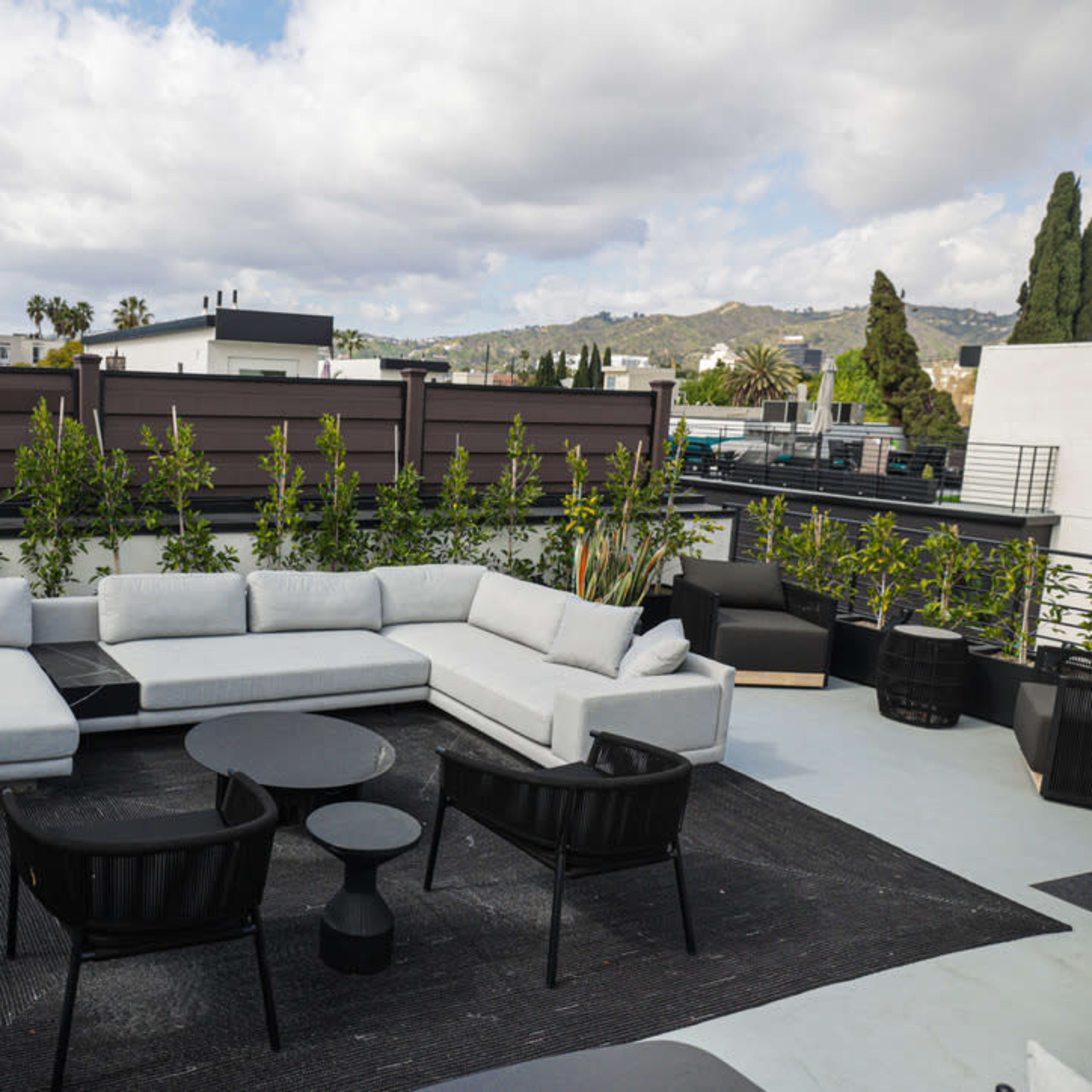 A modern rooftop lounge area featuring a sectional sofa, a circular black table, and two chairs, surrounded by greenery and a view of buildings and hills in the background.