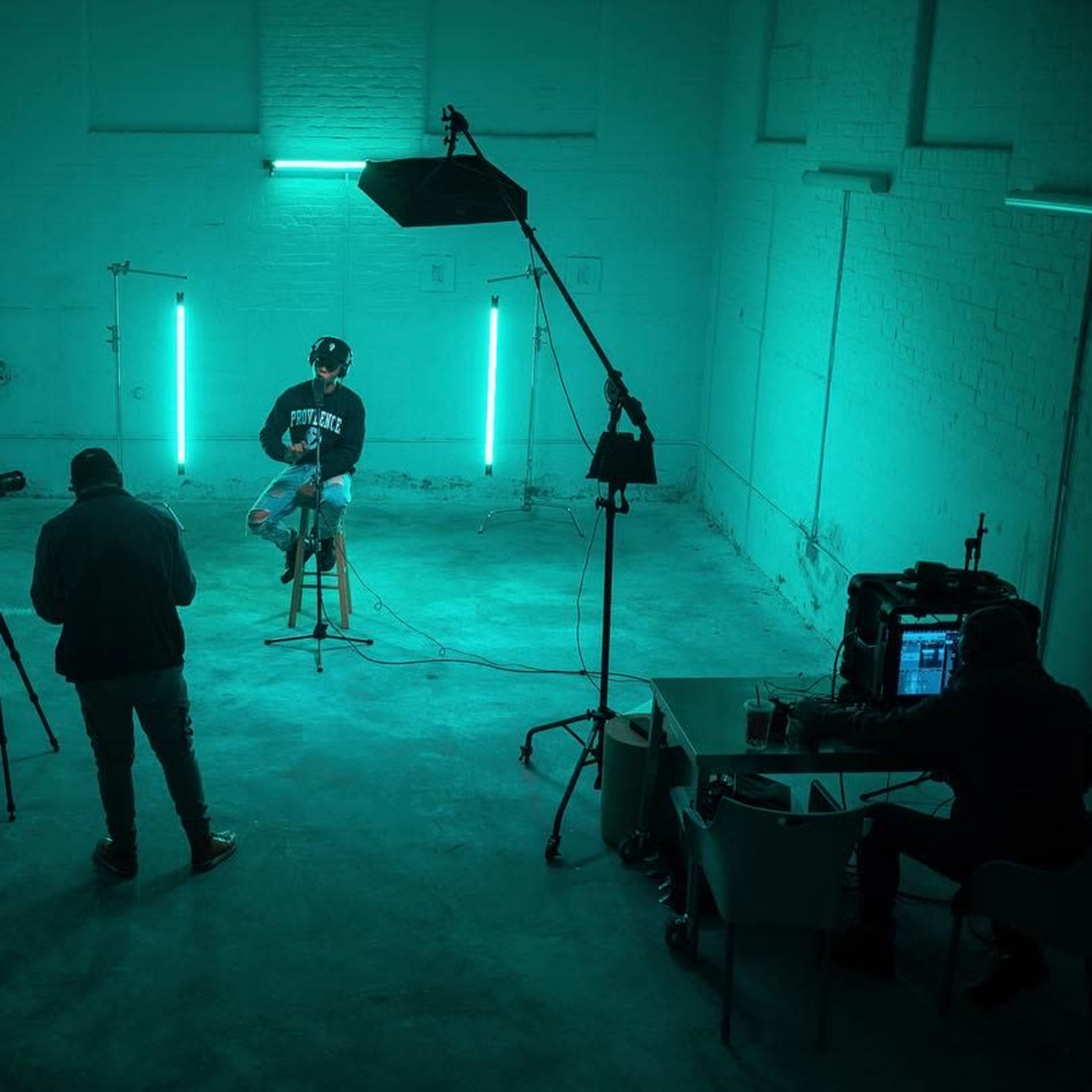 A person sits on a stool in a dimly lit room illuminated by teal lights, while two individuals operate cameras and a third monitors equipment at a desk.