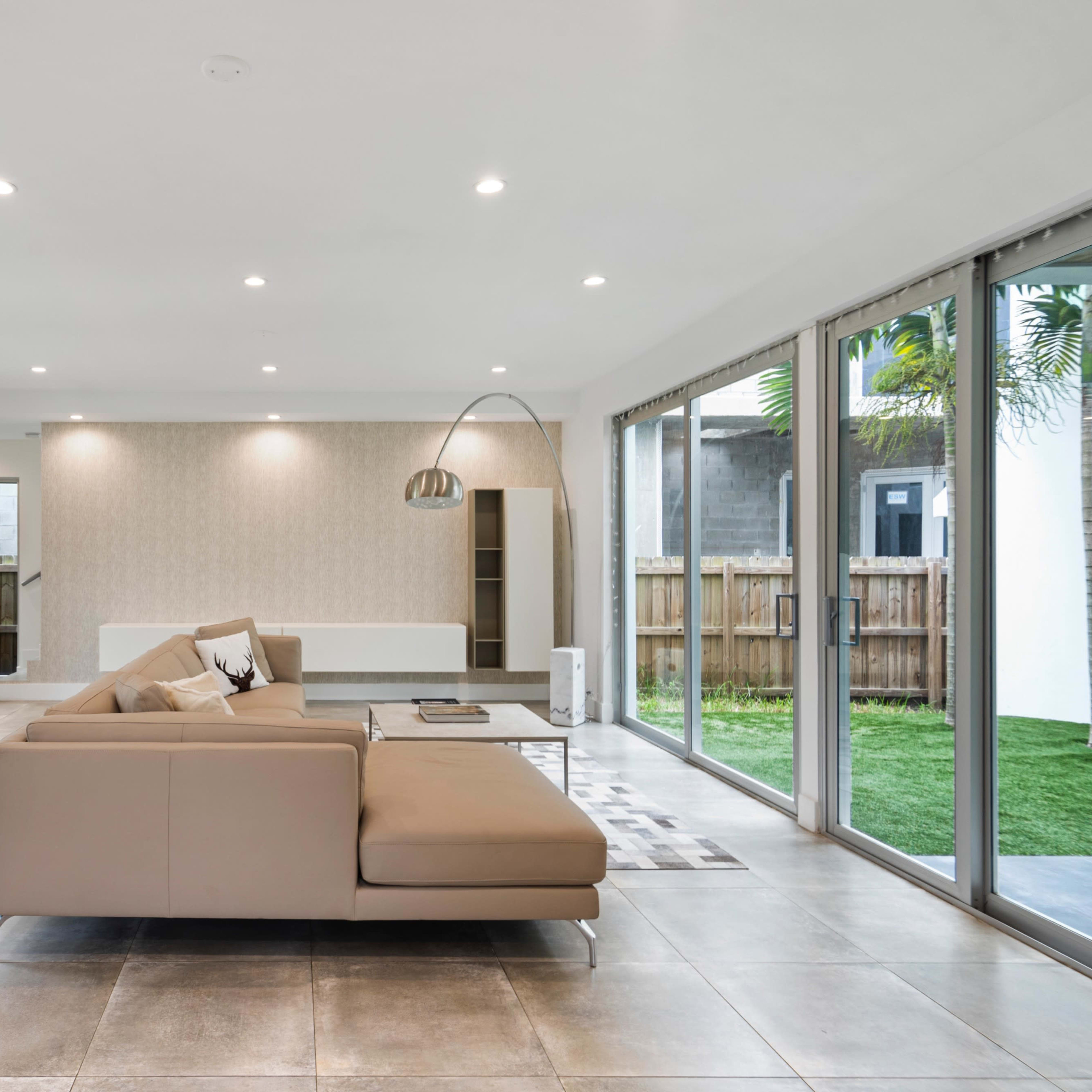 A modern living room with large glass doors, a beige sectional sofa, and a minimalist coffee table, surrounded by greenery.