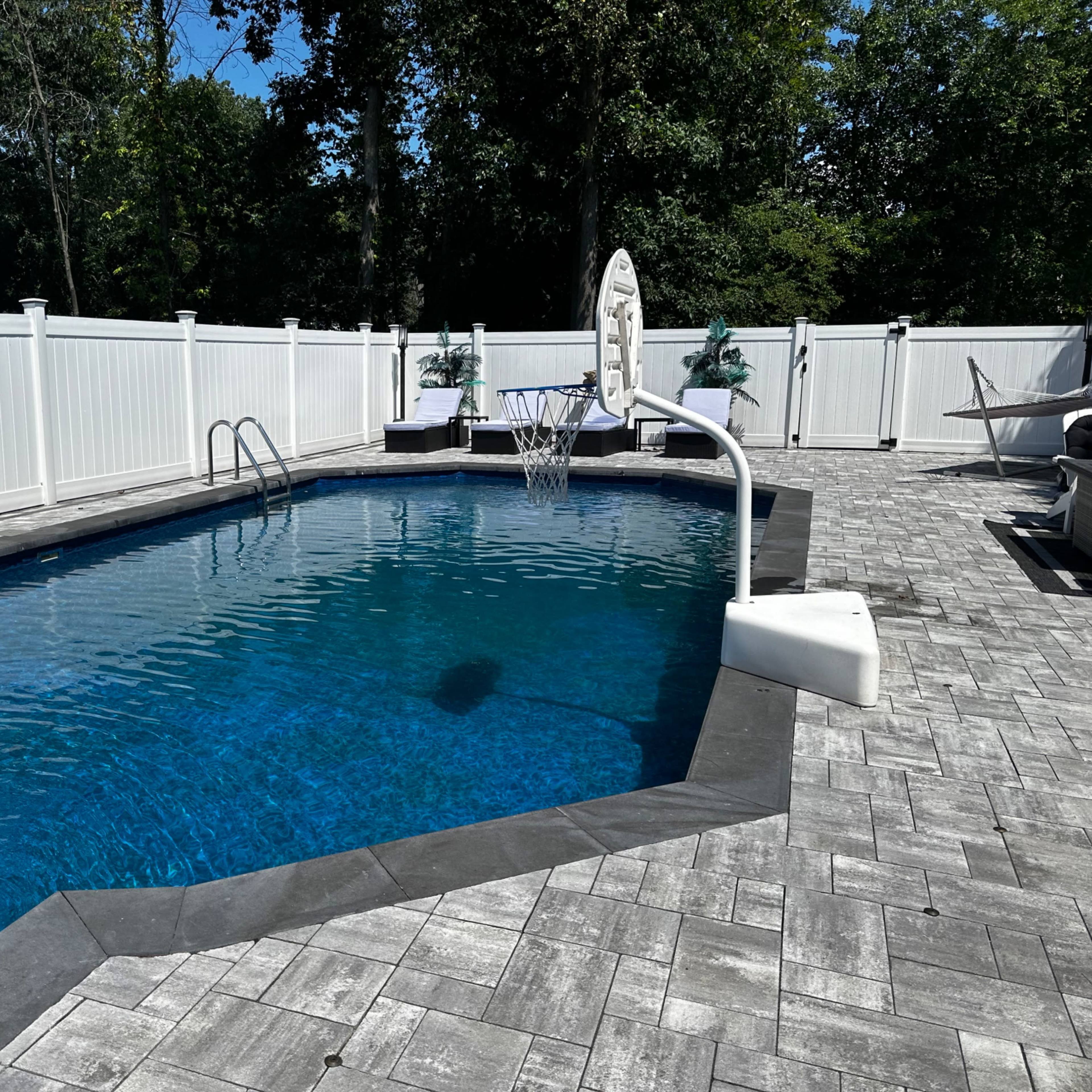 A blue swimming pool with a basketball hoop is situated on a stone patio surrounded by a white fence and trees.
