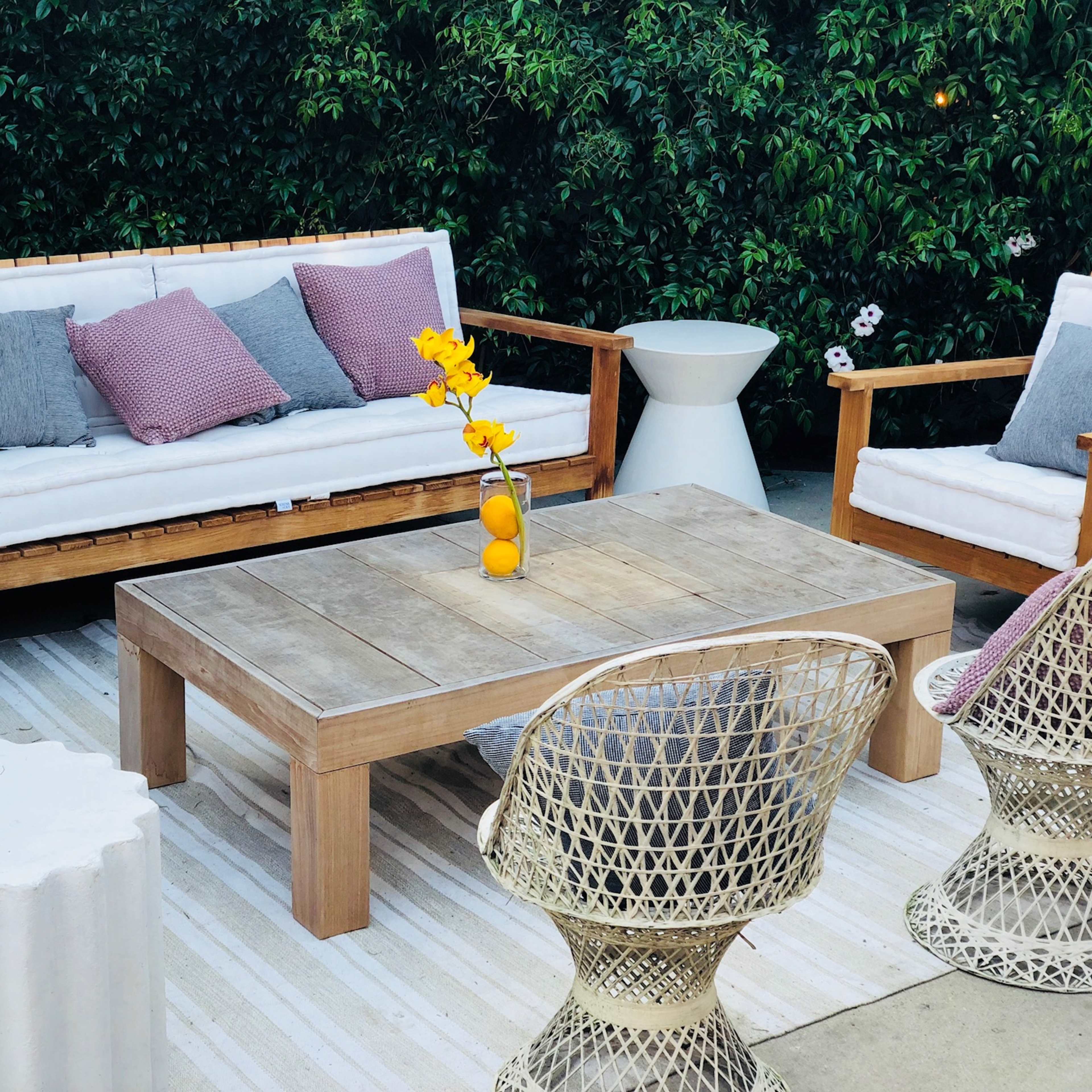A patio seating area features wooden chairs and a table, with cushions and a flower vase, surrounded by greenery.