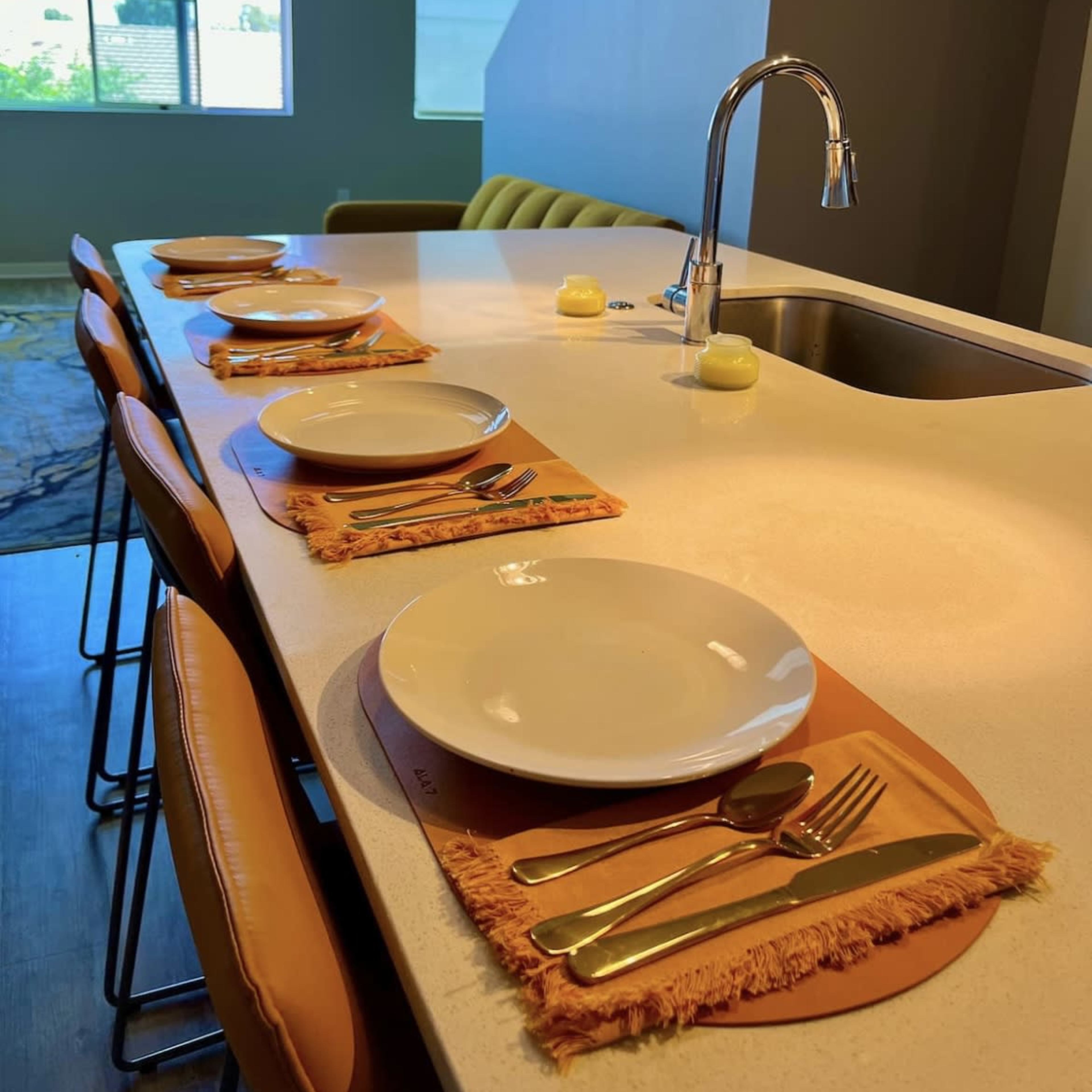 A neatly set kitchen island with four plates, utensils, and candles placed on orange placemats, all under natural light from a window.