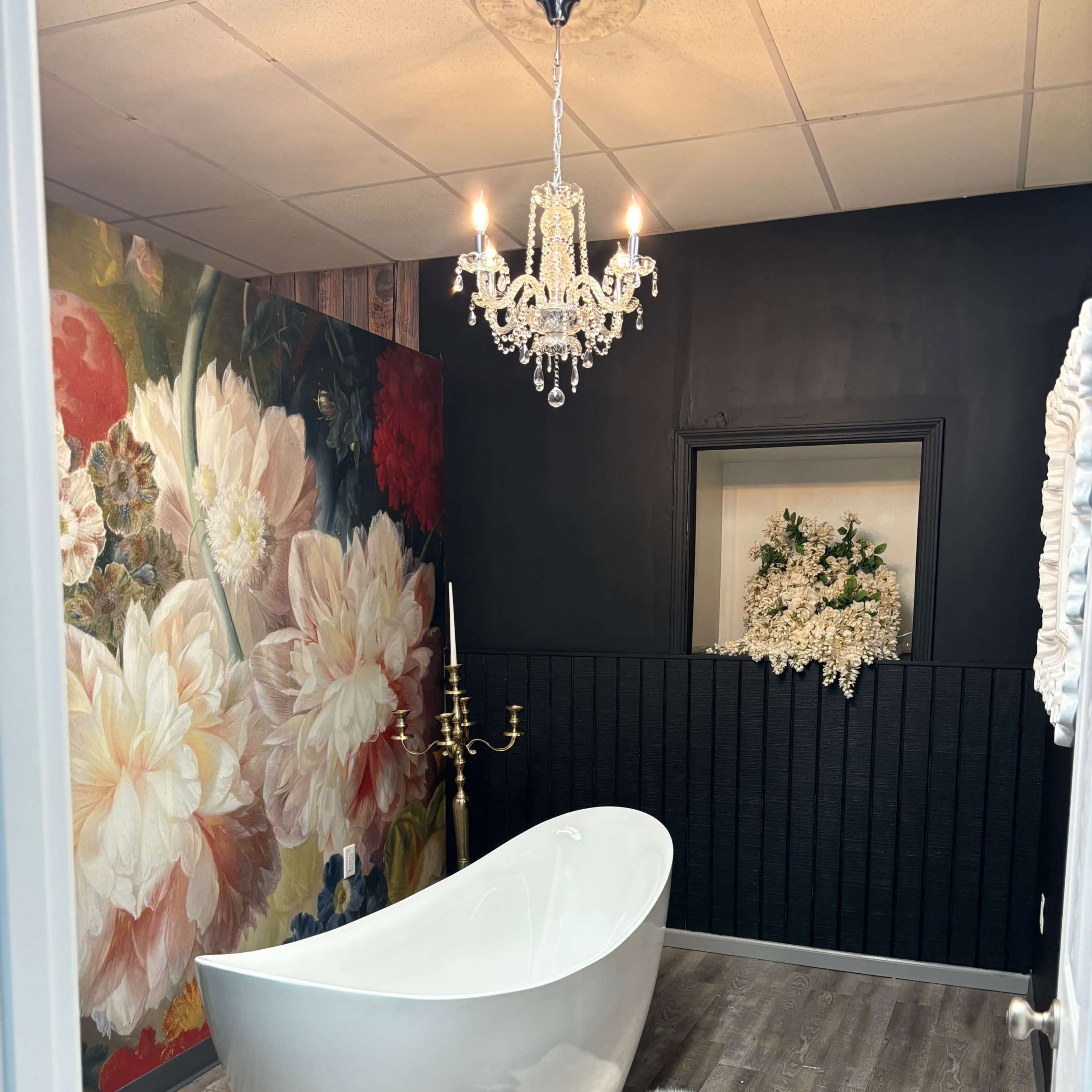 A modern bathroom featuring a white freestanding bathtub, a floral wall mural, and a chandelier hanging from the ceiling.