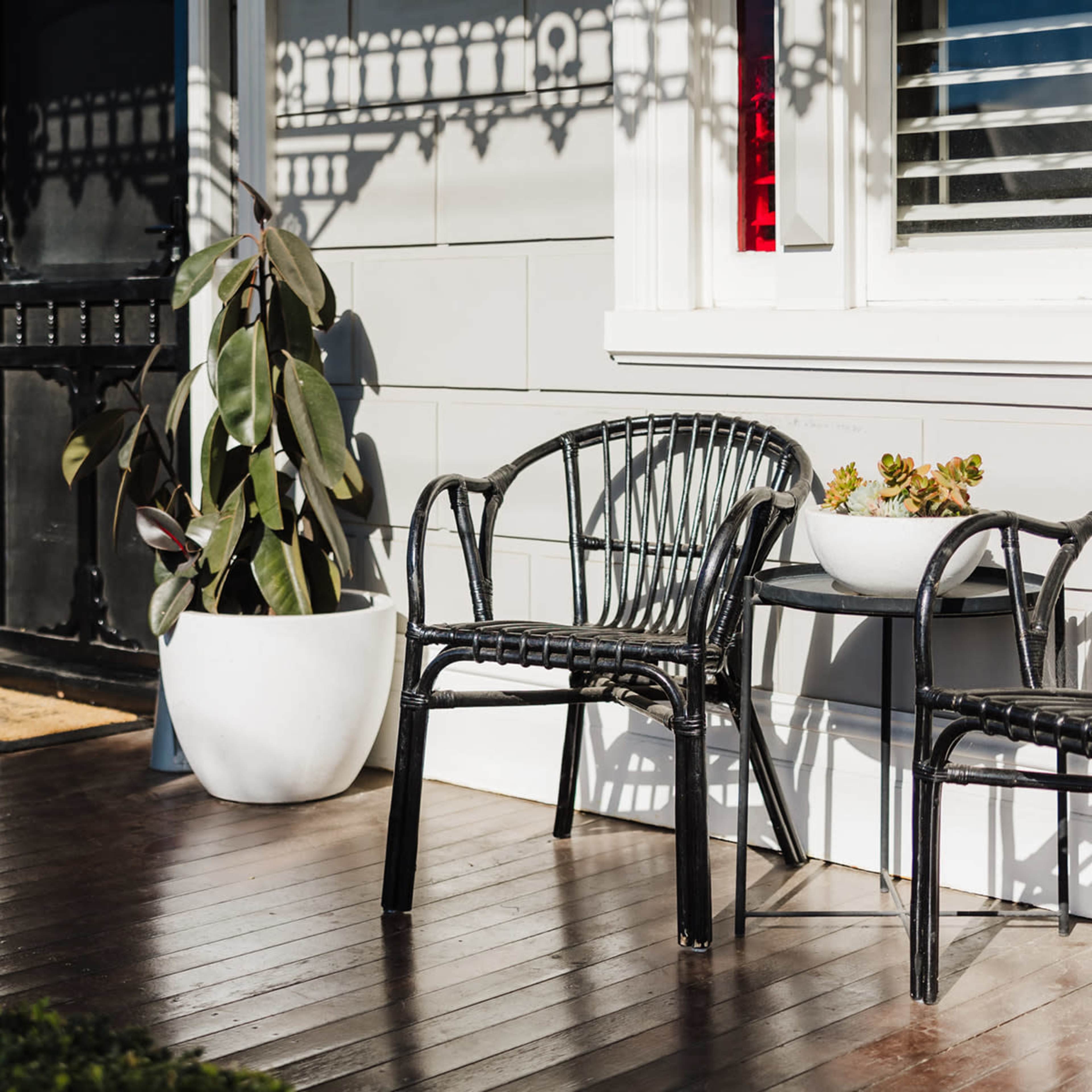 Two black wicker chairs and a small table sit on a wooden porch next to a large potted plant and a decorative bowl.