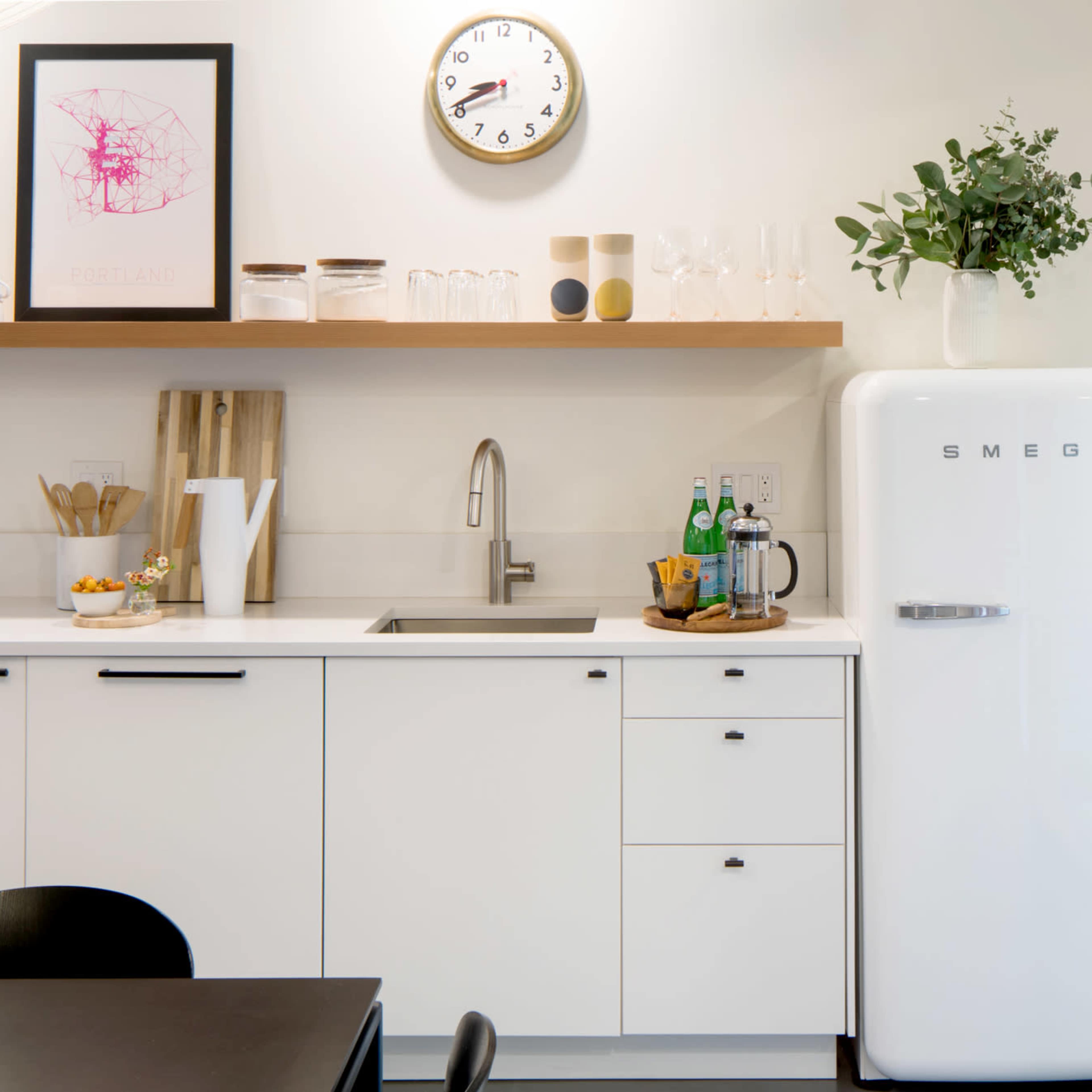 A modern kitchen features a white SMEG refrigerator, a sink, wooden shelves with glassware, and a clock on the wall, set against a minimalist backdrop.